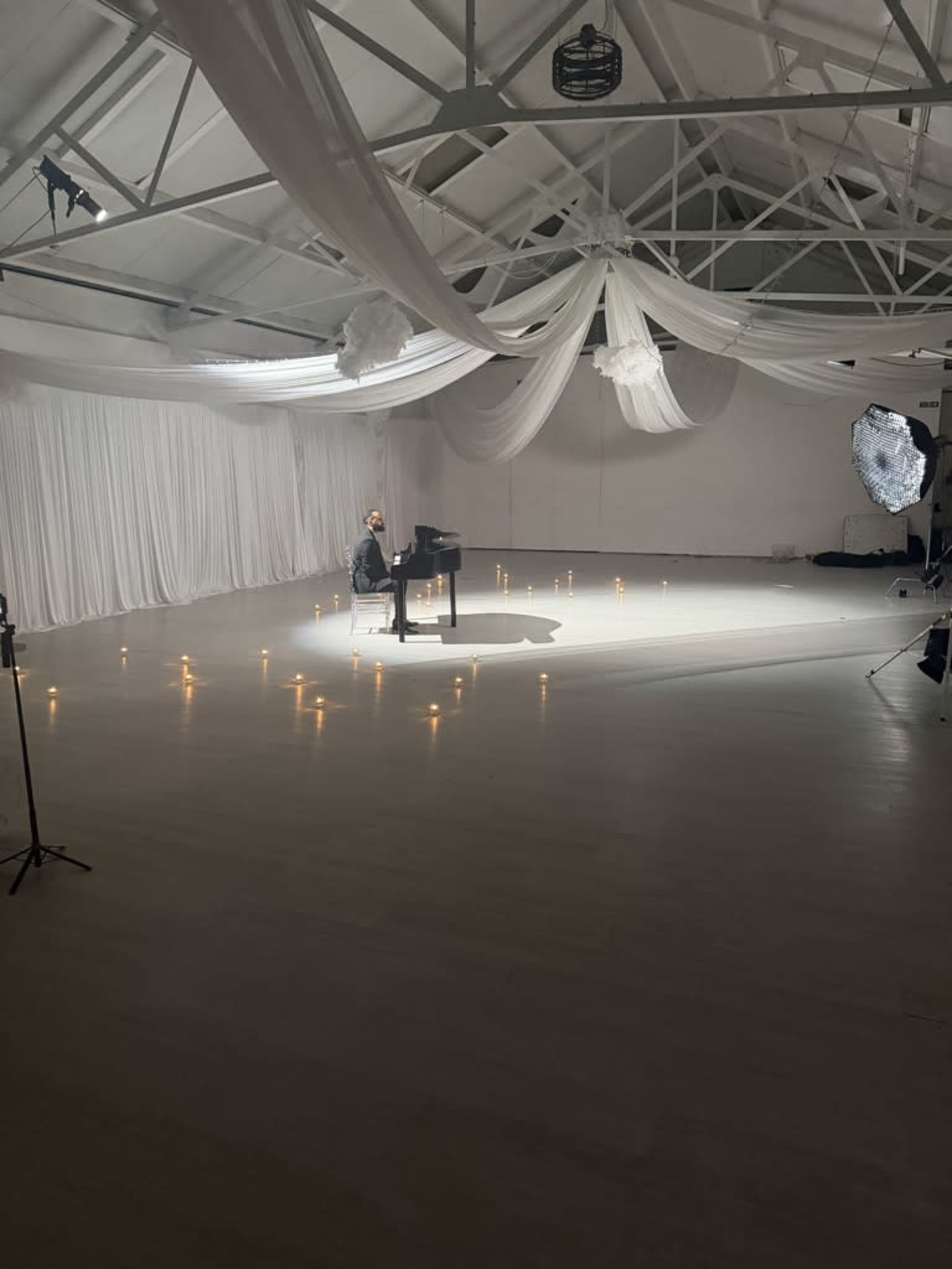 A person is playing a piano in a spacious, well-lit room with draped fabric and candles arranged on the floor.