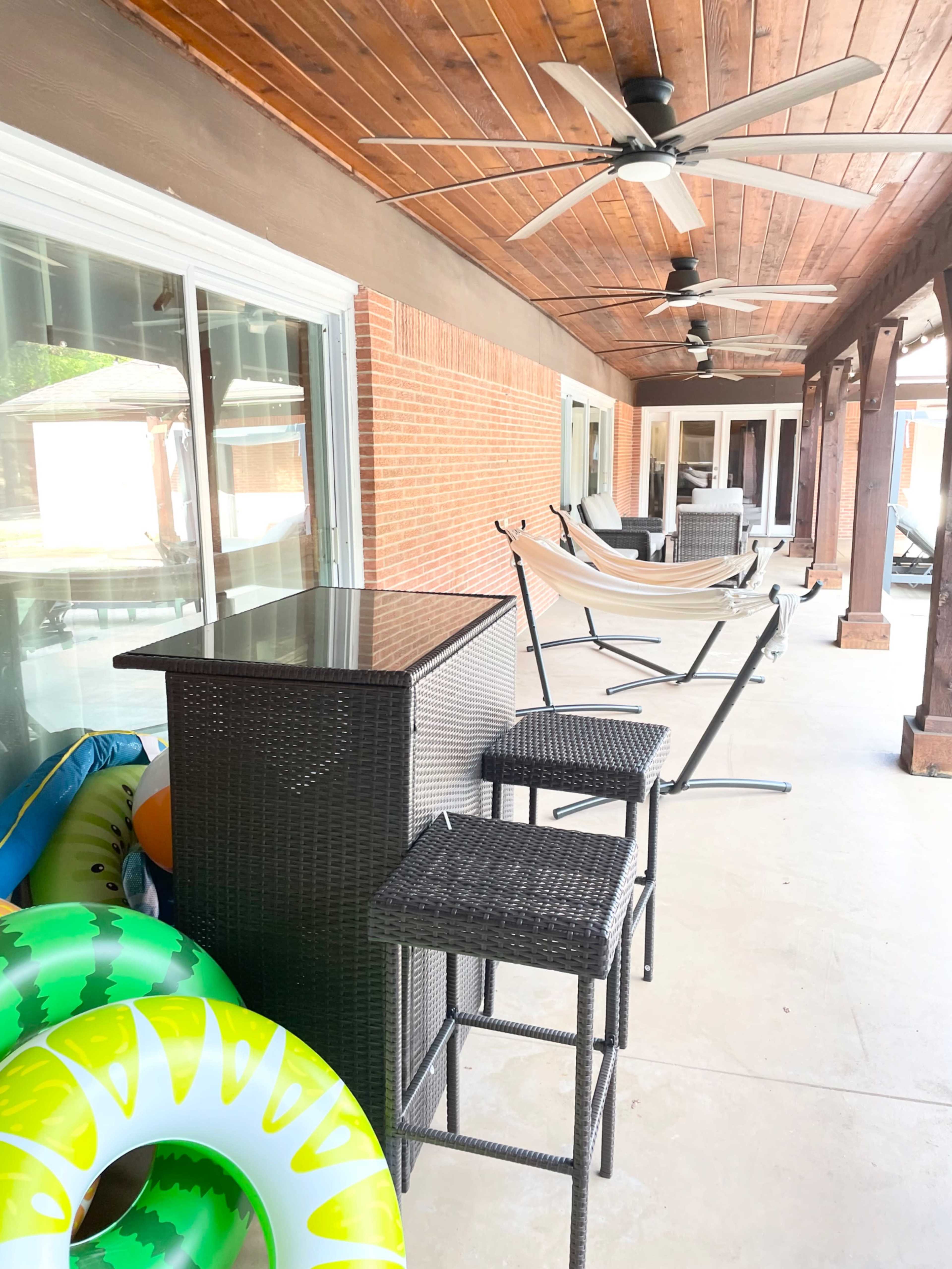 The image shows a covered outdoor patio area featuring a bar with two stools, a hammock, and colorful pool inflatables stacked nearby.