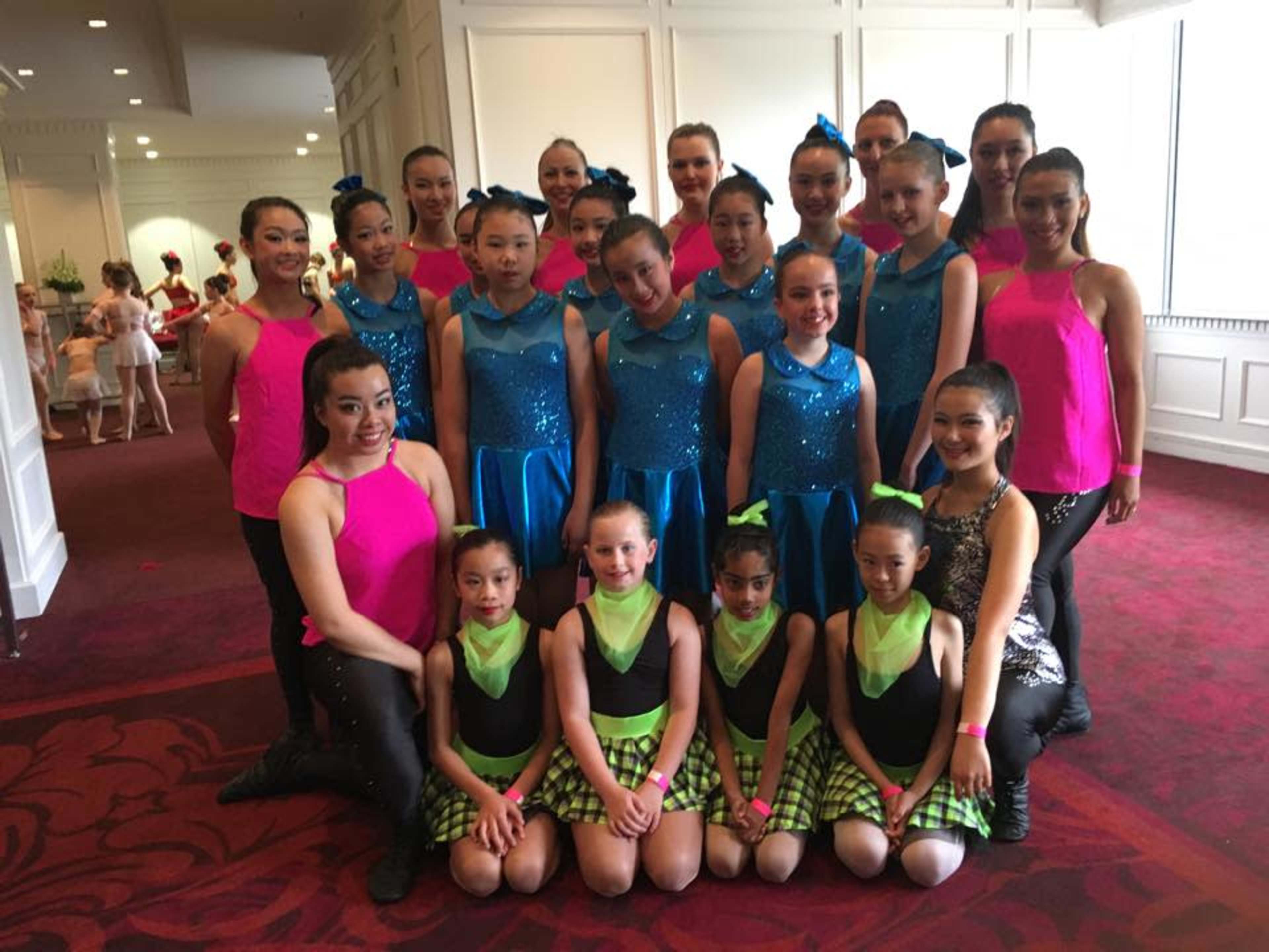 A group of young dancers in colorful costumes poses together in a well-lit interior space.