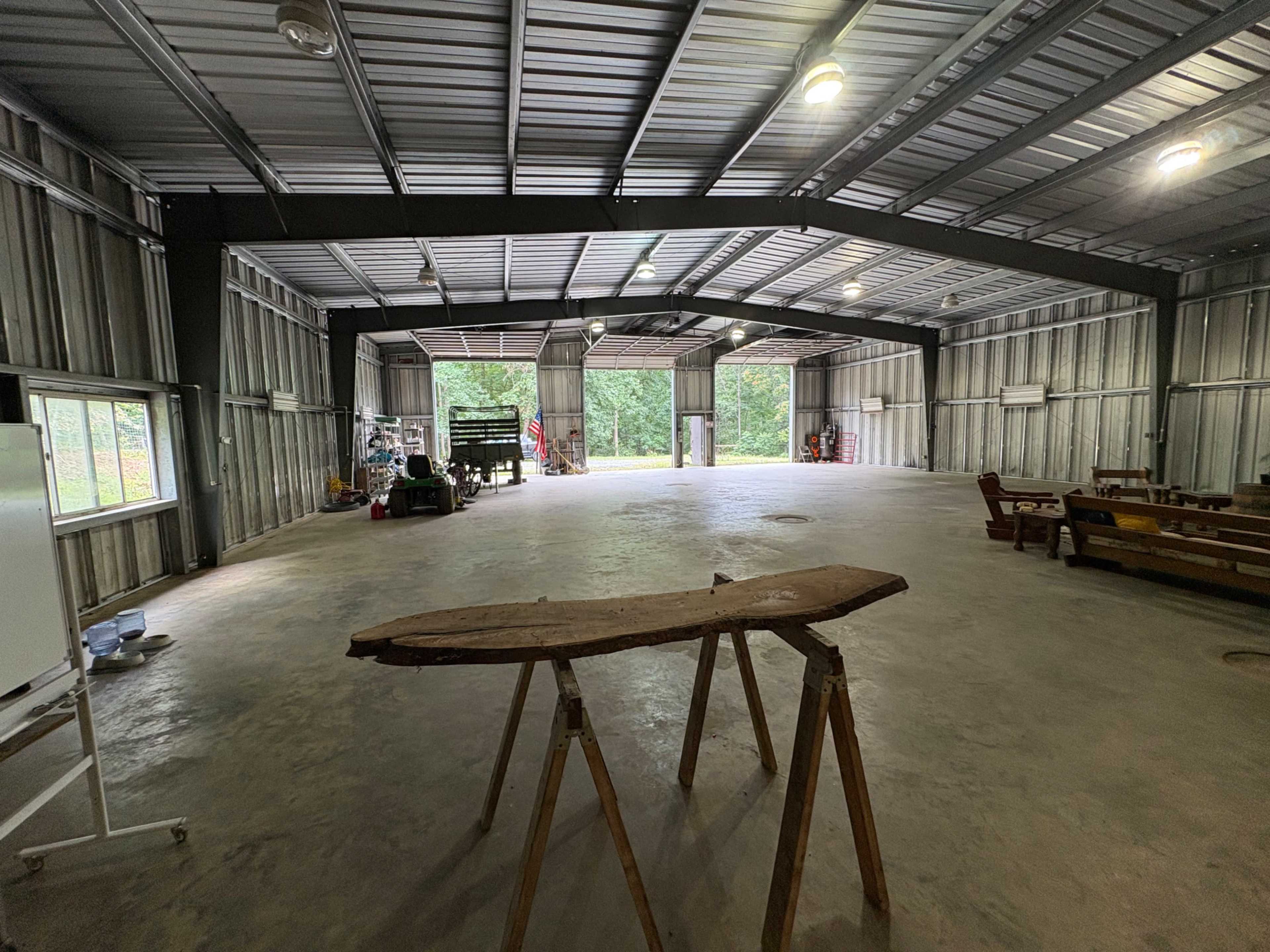 The image shows a spacious, empty industrial building with metal walls and a concrete floor, featuring wooden furniture and various tools in the background.