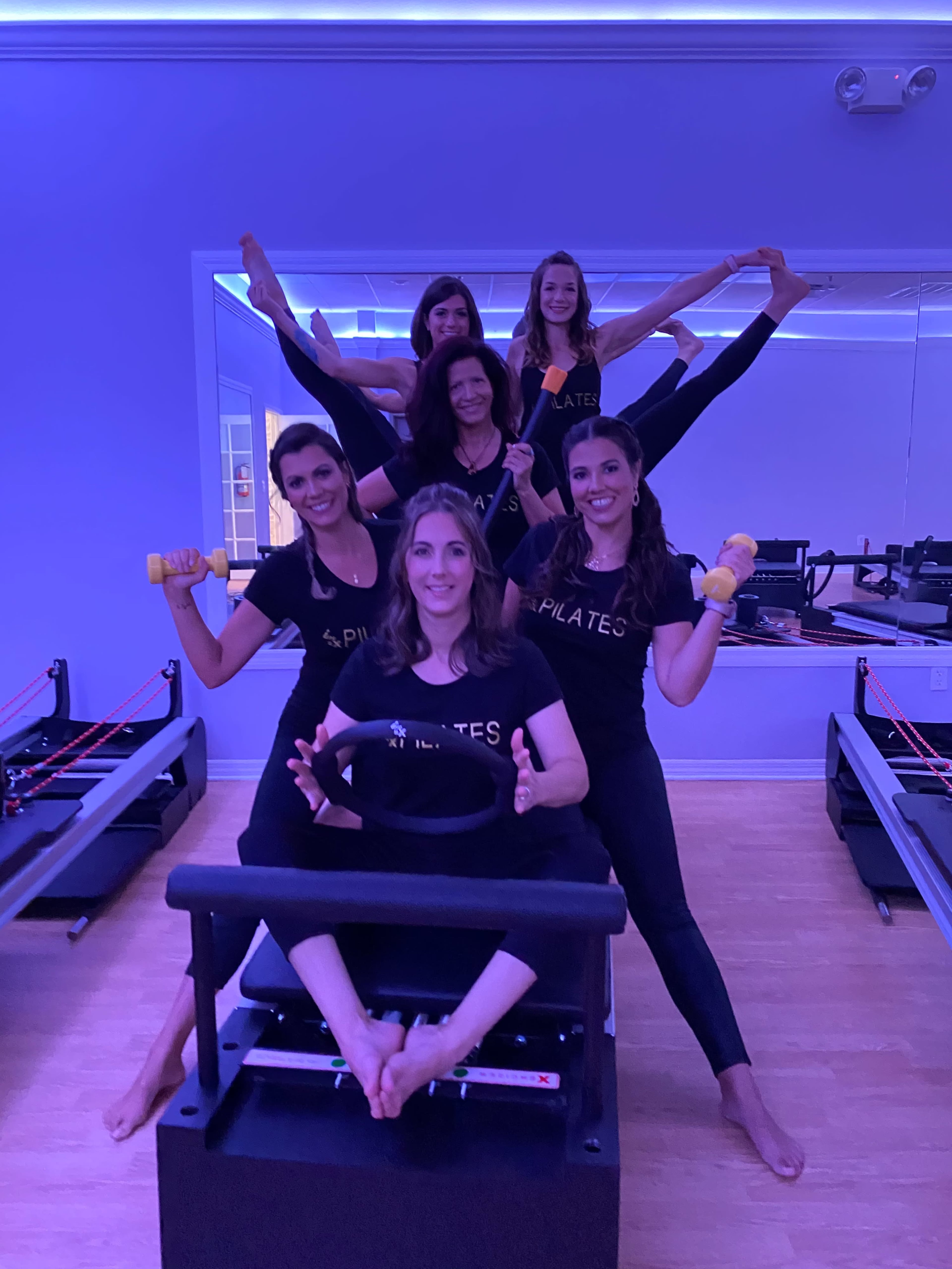 A group of six women in a Pilates studio, performing various exercises using equipment and props under purple lighting.