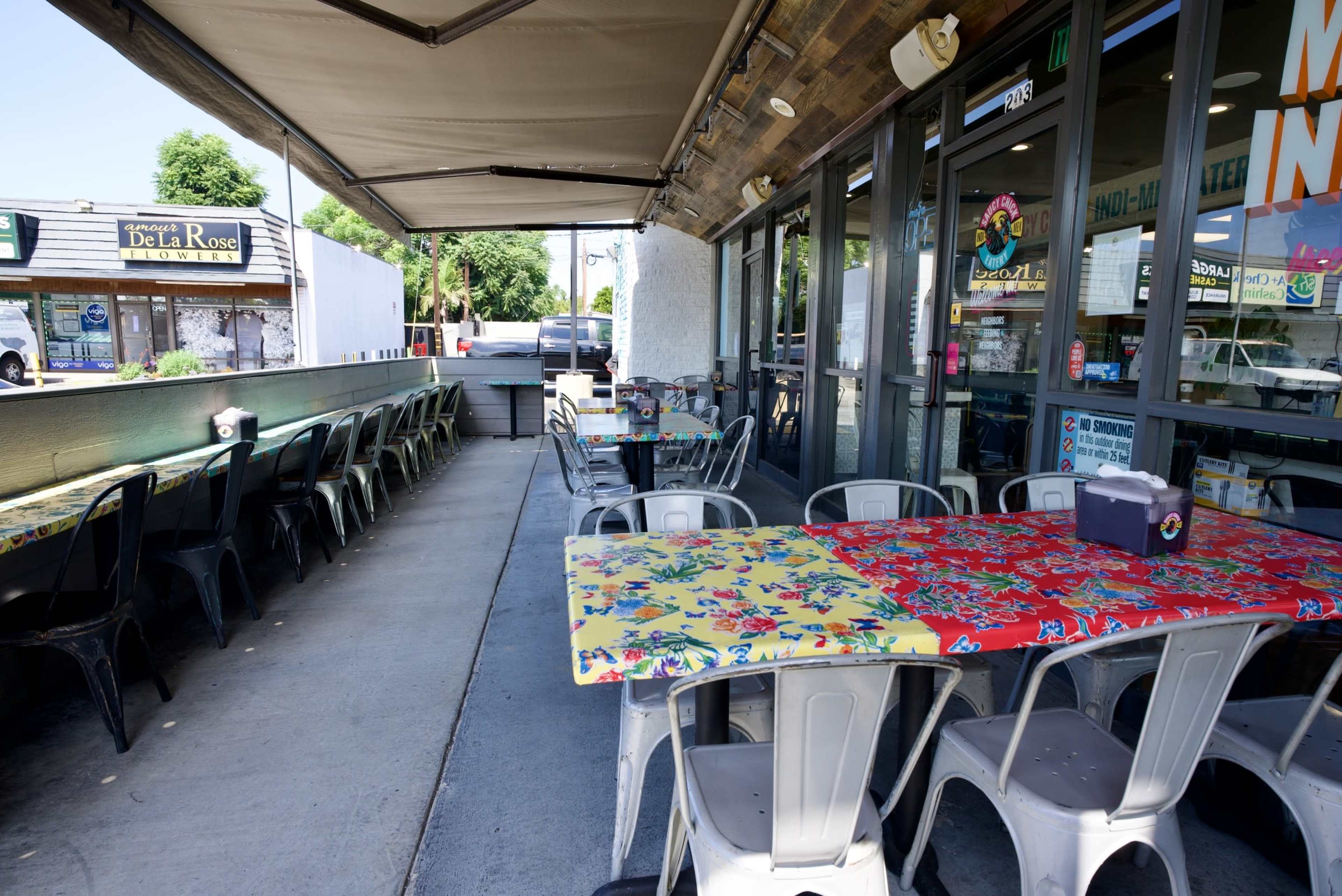The image shows a covered outdoor dining area of a restaurant with colorful tables and metal chairs arranged neatly.