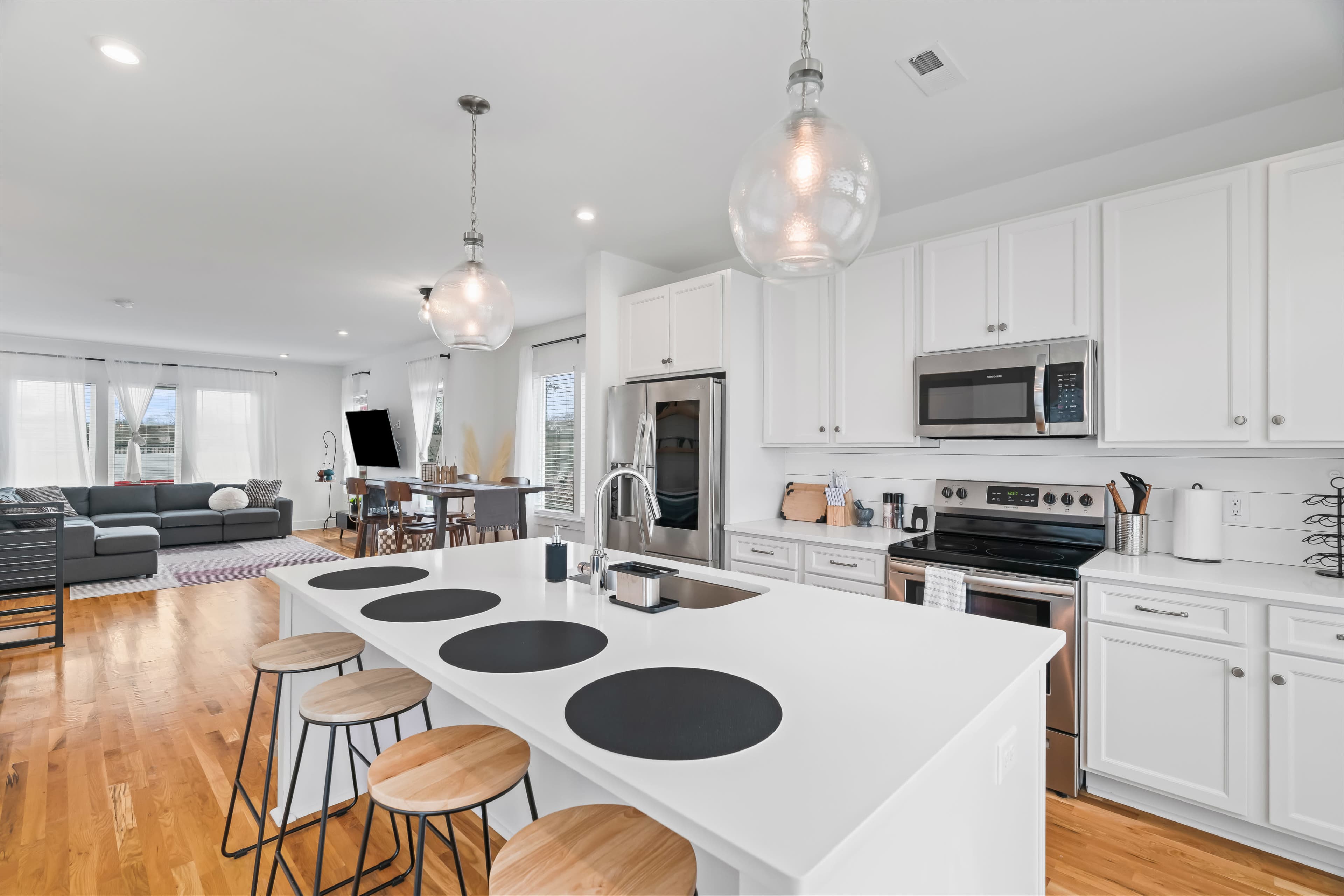 A modern kitchen features a large white island with black placemats, wooden stools, stainless steel appliances, and an open layout that connects to a living area.