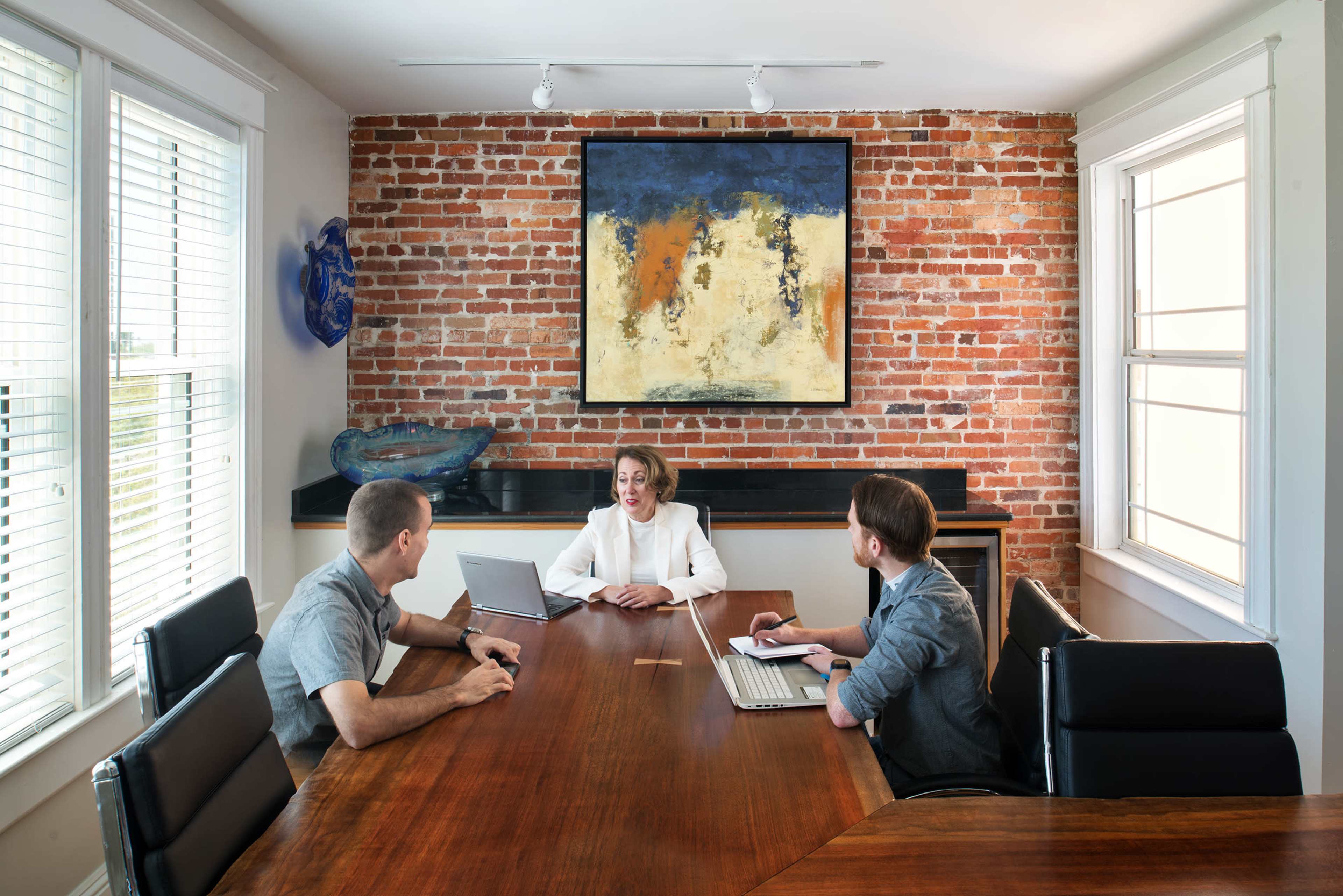 Three people are seated around a wooden conference table in a well-lit room with exposed brick walls and a large painting.