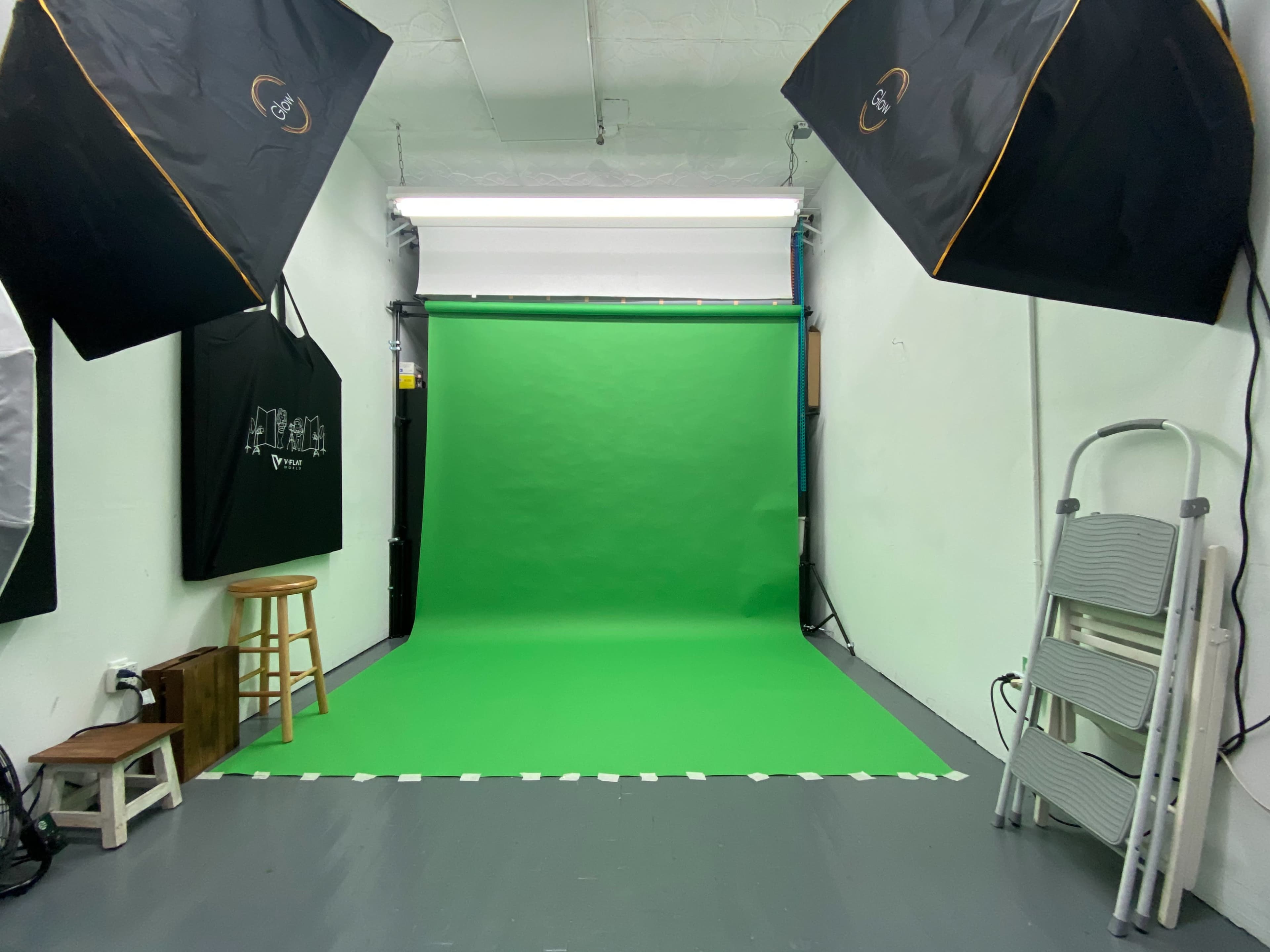 A photography studio with a green screen backdrop, two softbox lights, and various equipment including stools and a step ladder.