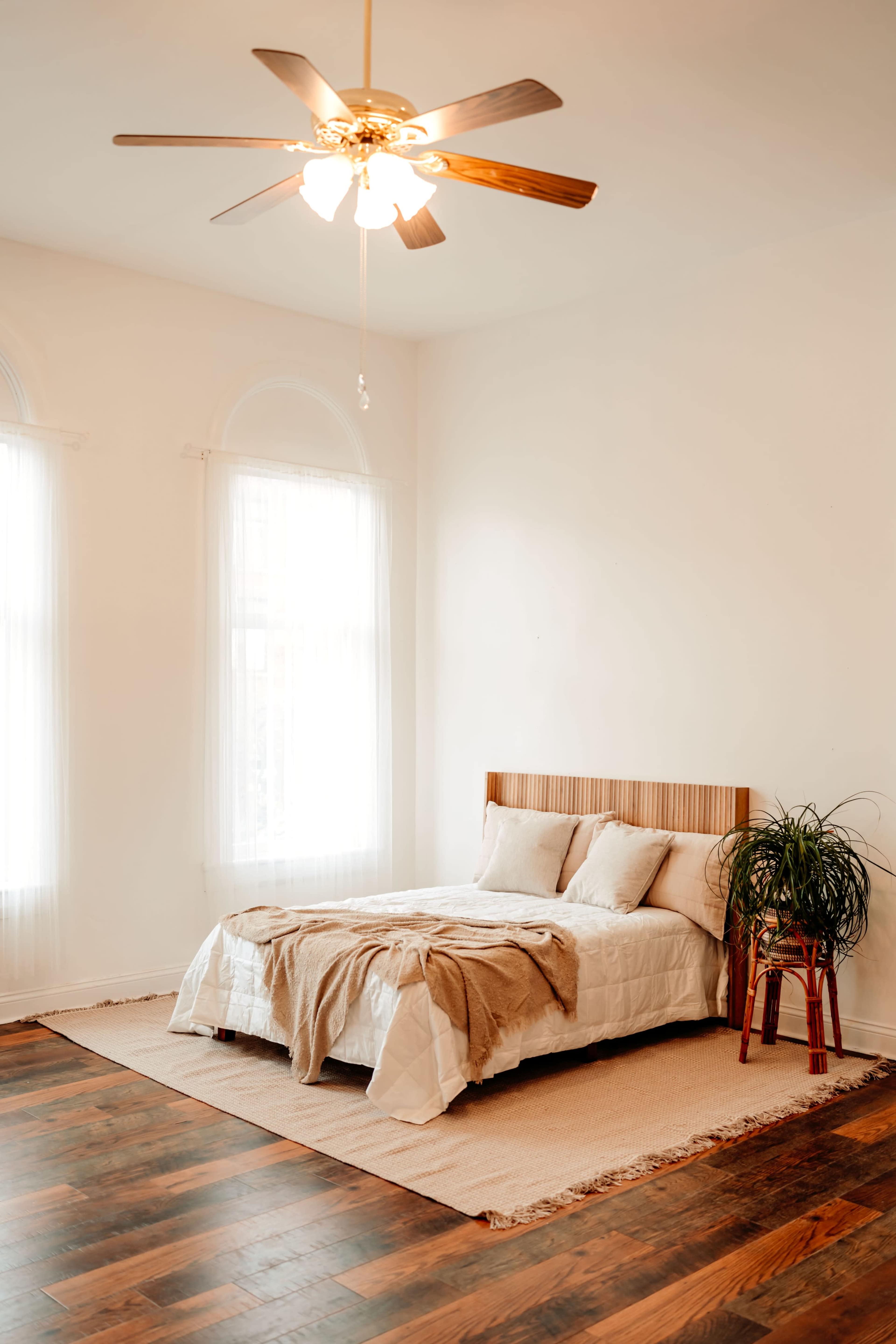 A neatly arranged bedroom features a bed with a light-colored comforter, two pillows, and a potted plant beside it, beneath a ceiling fan.
