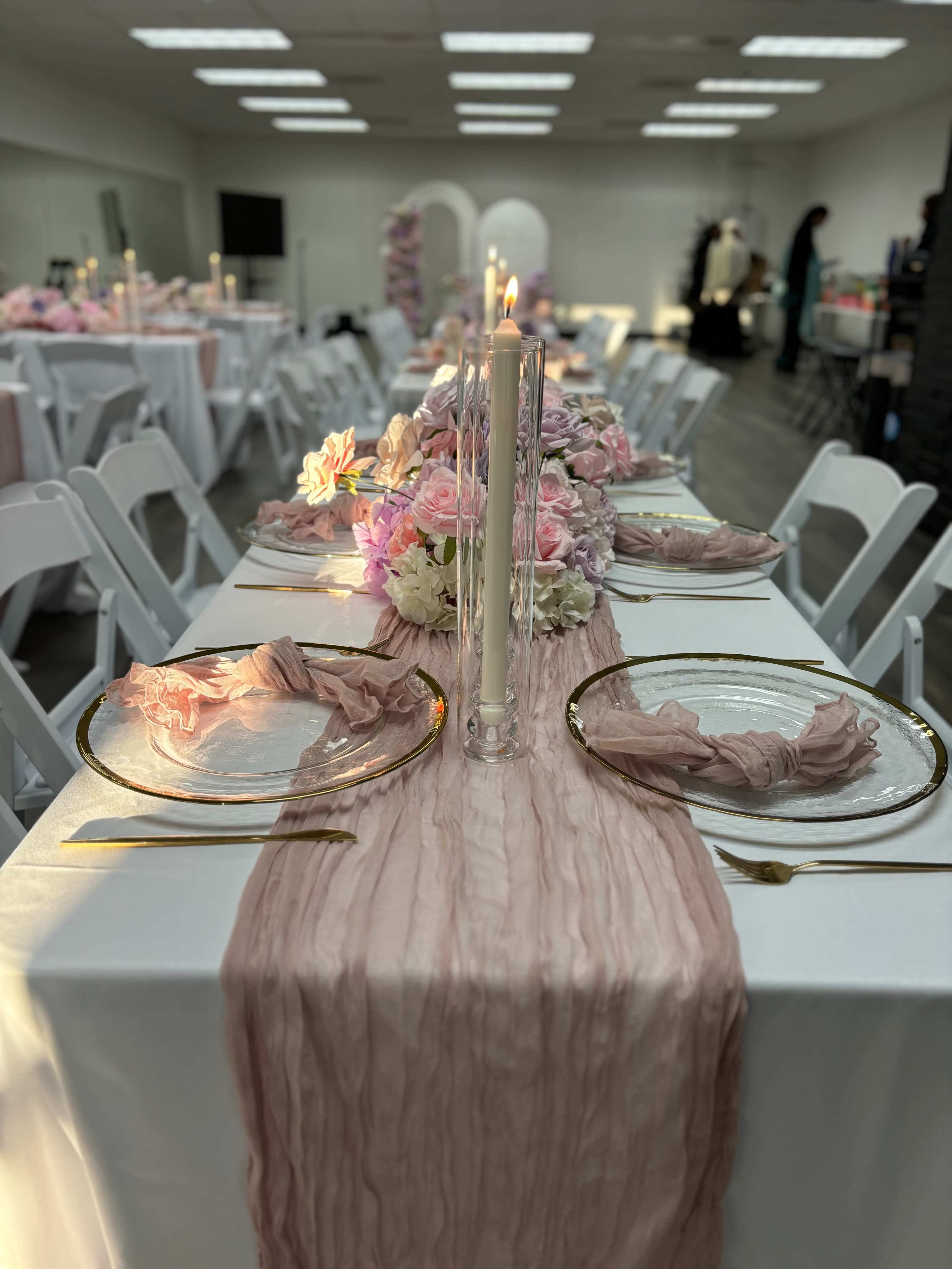 A long table is set with white linens, elegant glassware, tall candles, and floral arrangements in a well-lit event space.