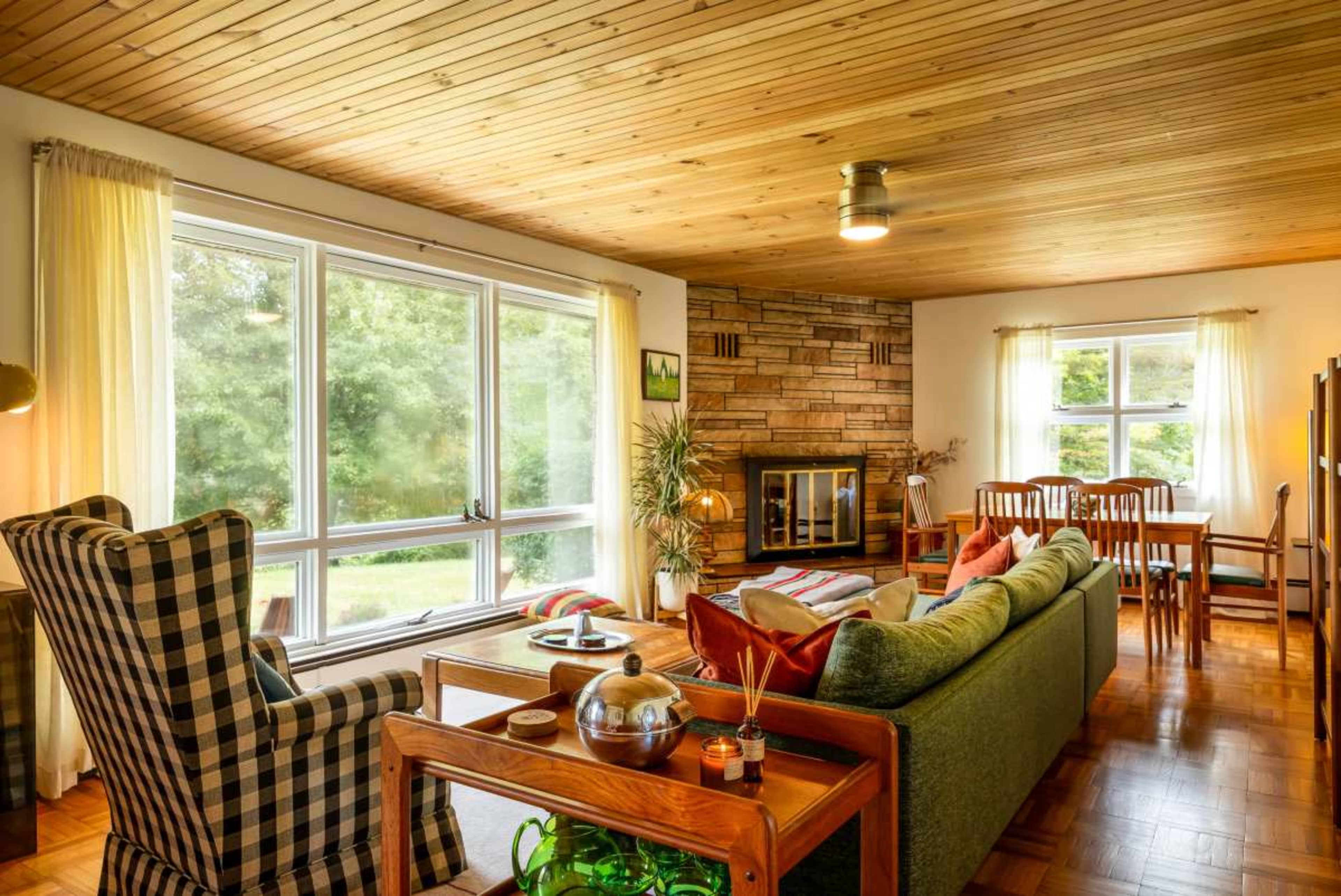 The image shows a cozy living room featuring a green sofa, a checkered armchair, wooden furniture, and large windows allowing natural light to fill the space.