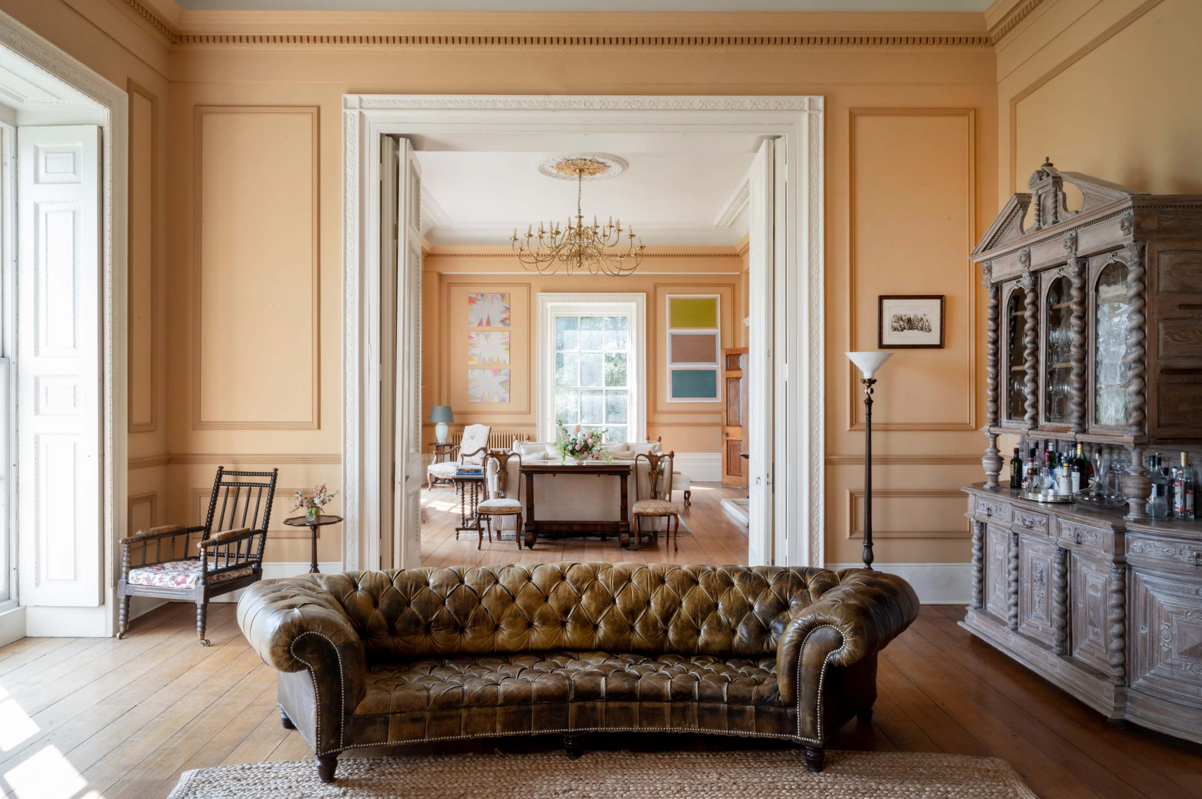 The image shows a spacious, elegantly furnished room with a brown tufted sofa in the foreground and a dining area visible through an open doorway in the background.