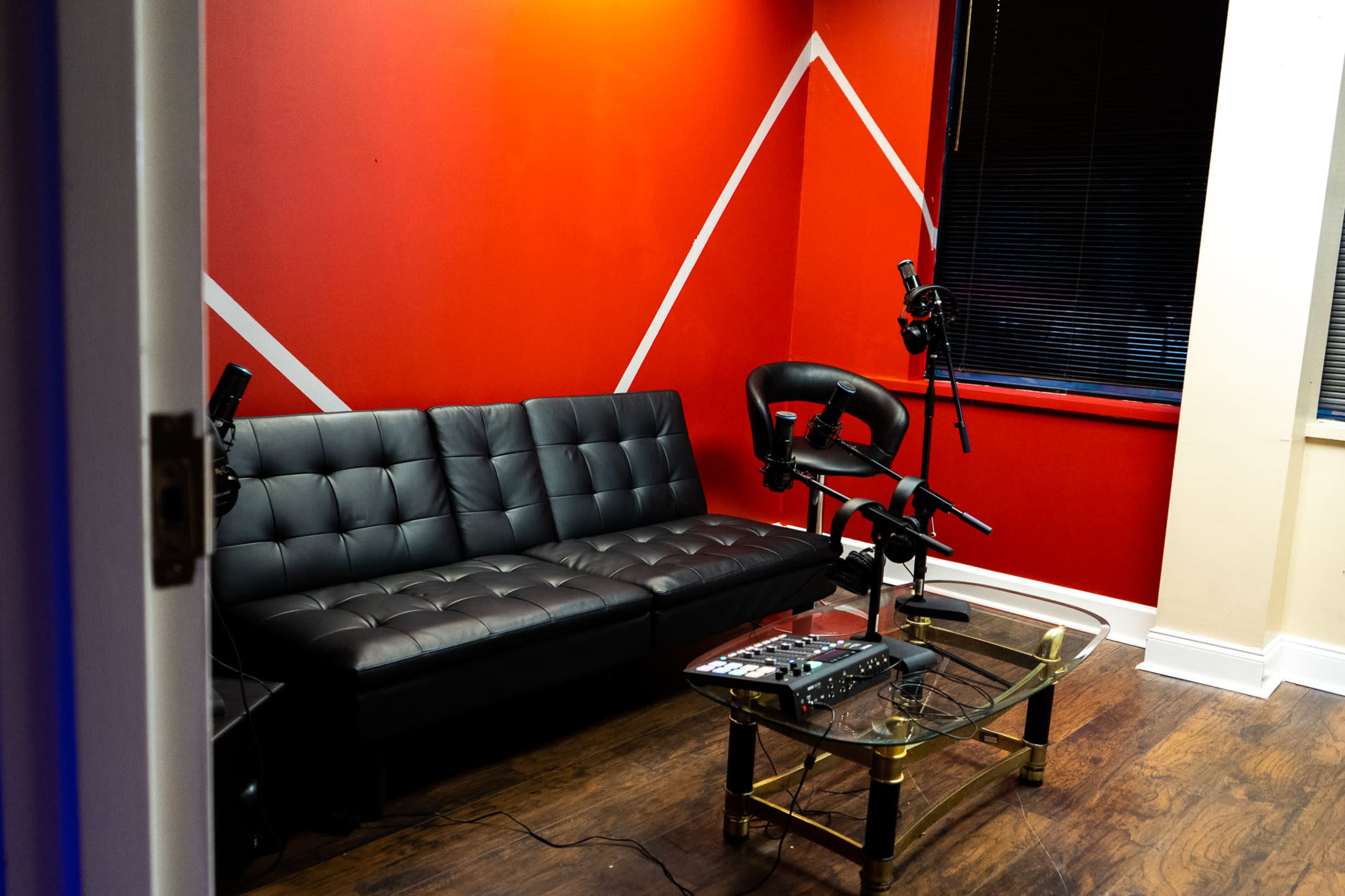 The image shows a small recording studio with a black couch, a glass coffee table, and two microphones positioned on stands against a red wall.