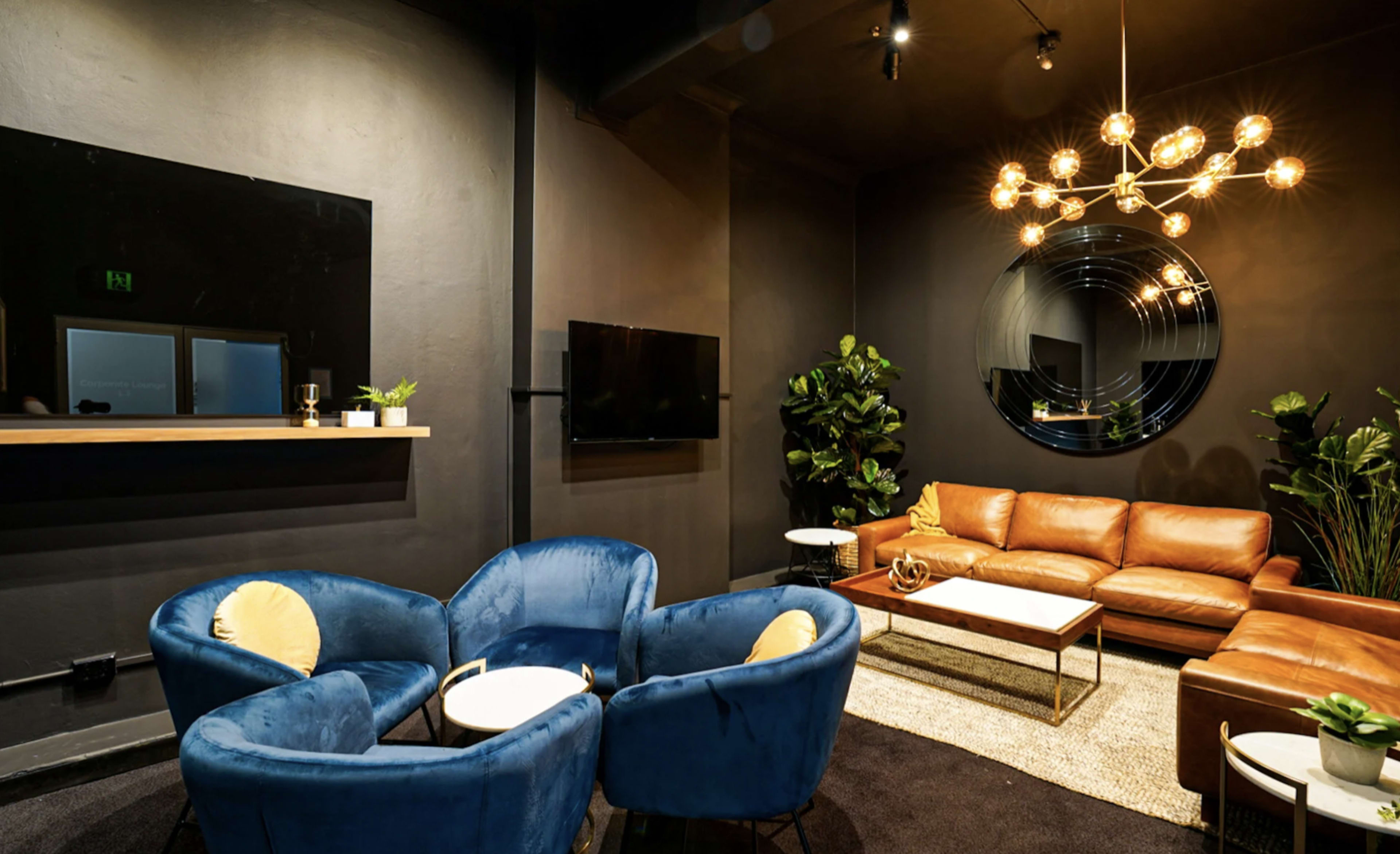 The image shows a modern lounge area with a brown leather sofa, blue velvet chairs, and a round mirror on a dark wall, complemented by greenery and stylish lighting.