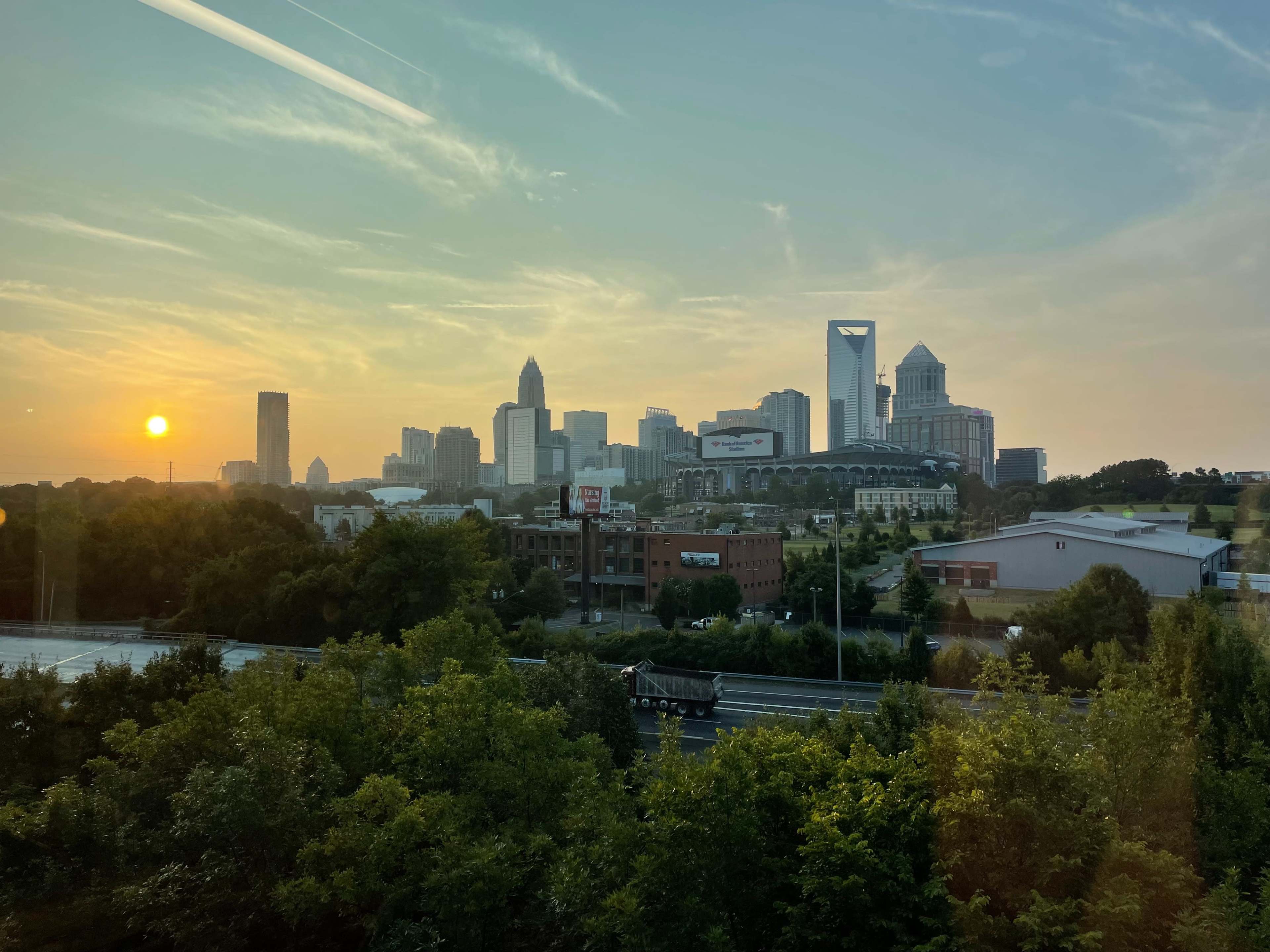 The image shows a sunset over a city skyline, with tall buildings in the background and a highway lined with trees in the foreground.