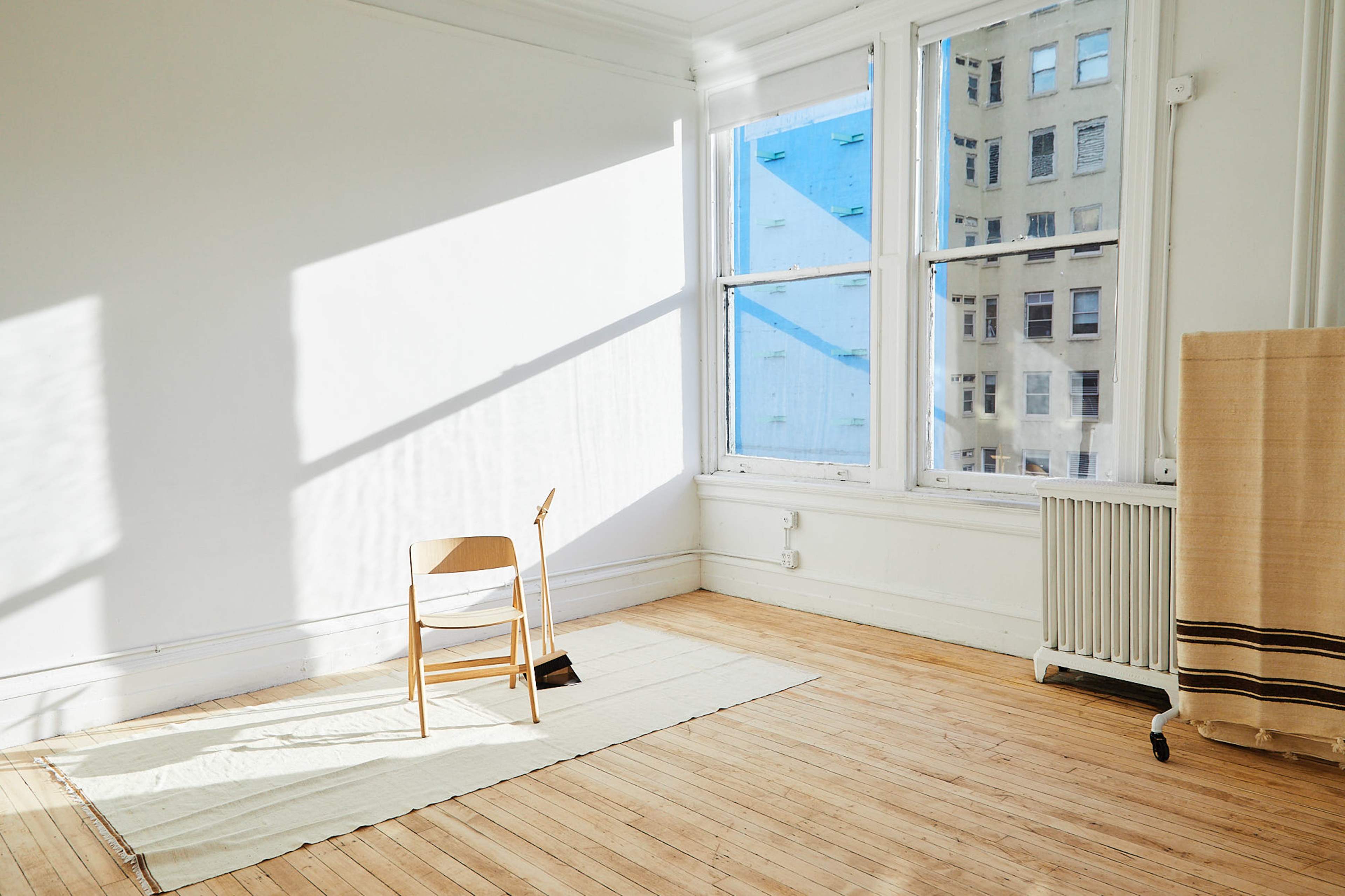 A wooden chair is positioned on a light-colored rug in a sunlit, empty room with large windows revealing a cityscape outside.