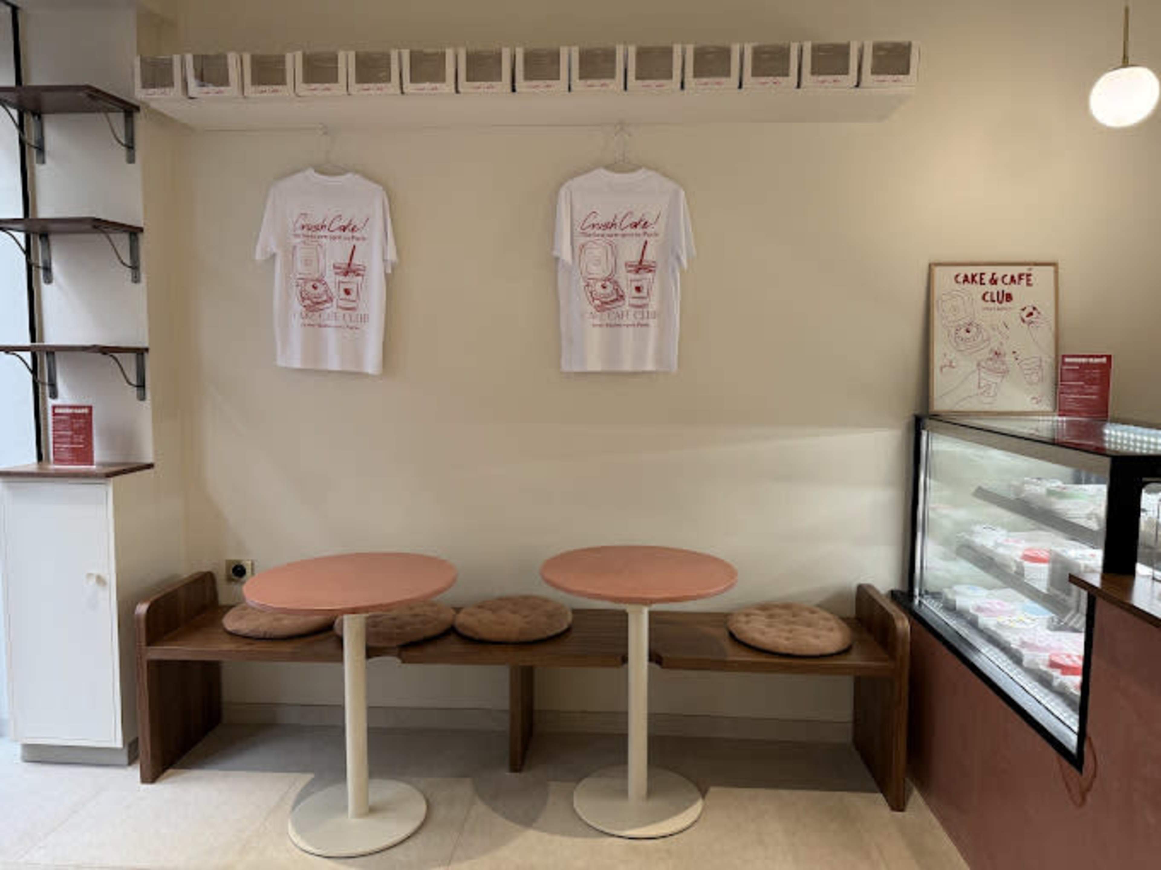 The image shows a cozy café interior featuring two round tables with cushioned benches, decorated with two hanging shirts and a display case filled with pastries.