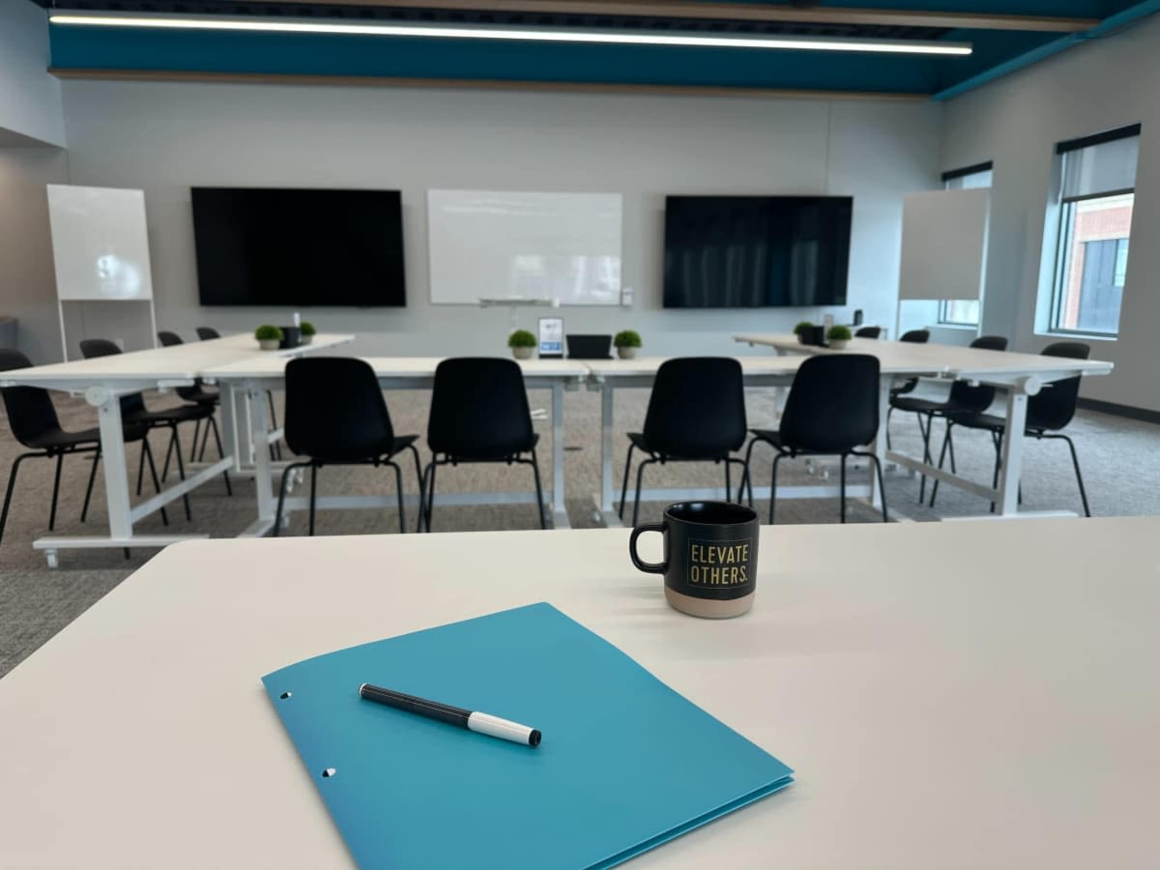 A spacious conference room features a large table with a blue folder and pen in the foreground, while multiple black chairs and display screens are arranged in the background.