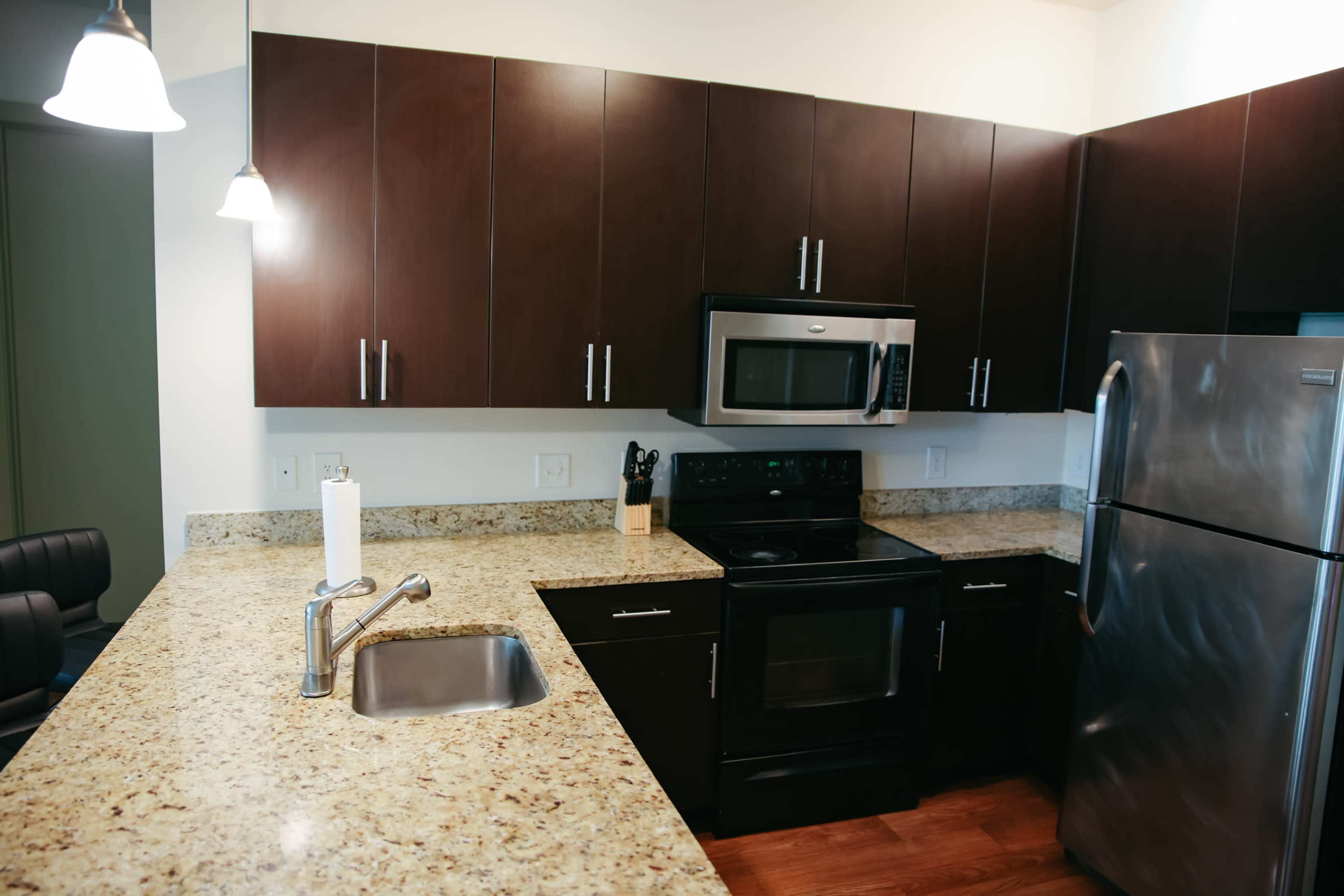 The image shows a modern kitchen with dark cabinets, a granite countertop, stainless steel appliances, and a single basin sink.