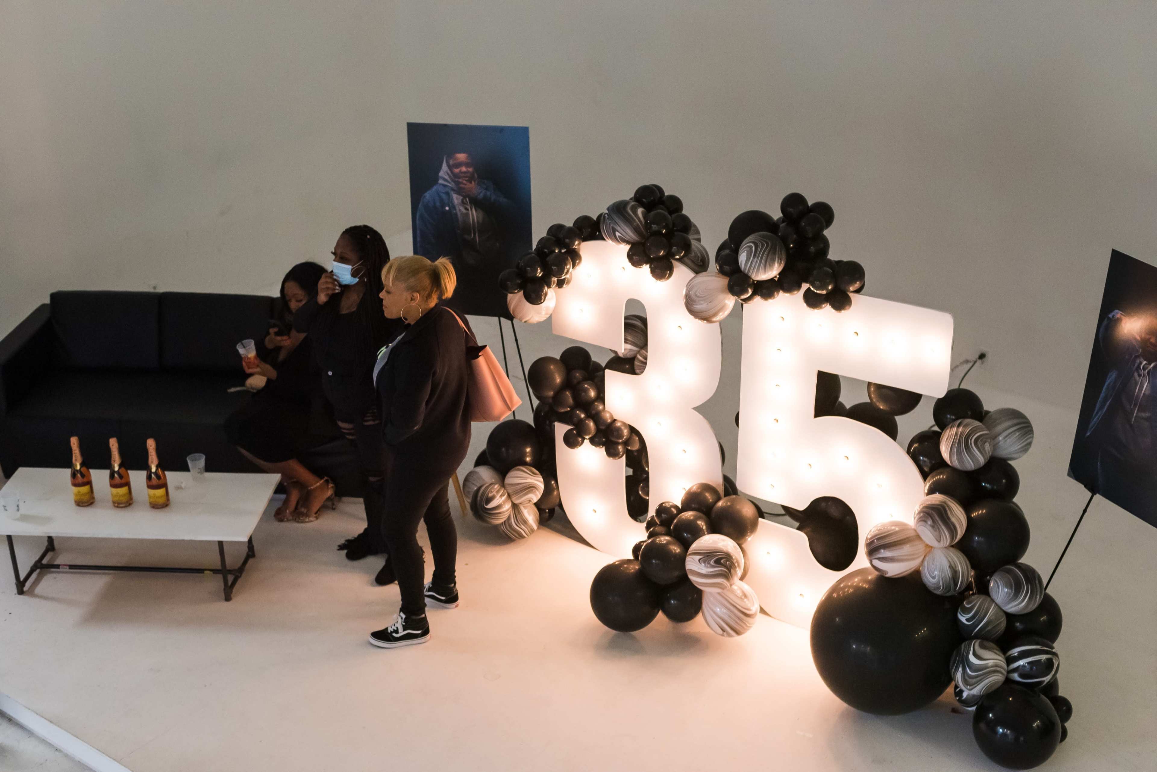 A group of three people stands next to a large illuminated "35" display decorated with black and silver balloons, while bottles are arranged on a nearby table.