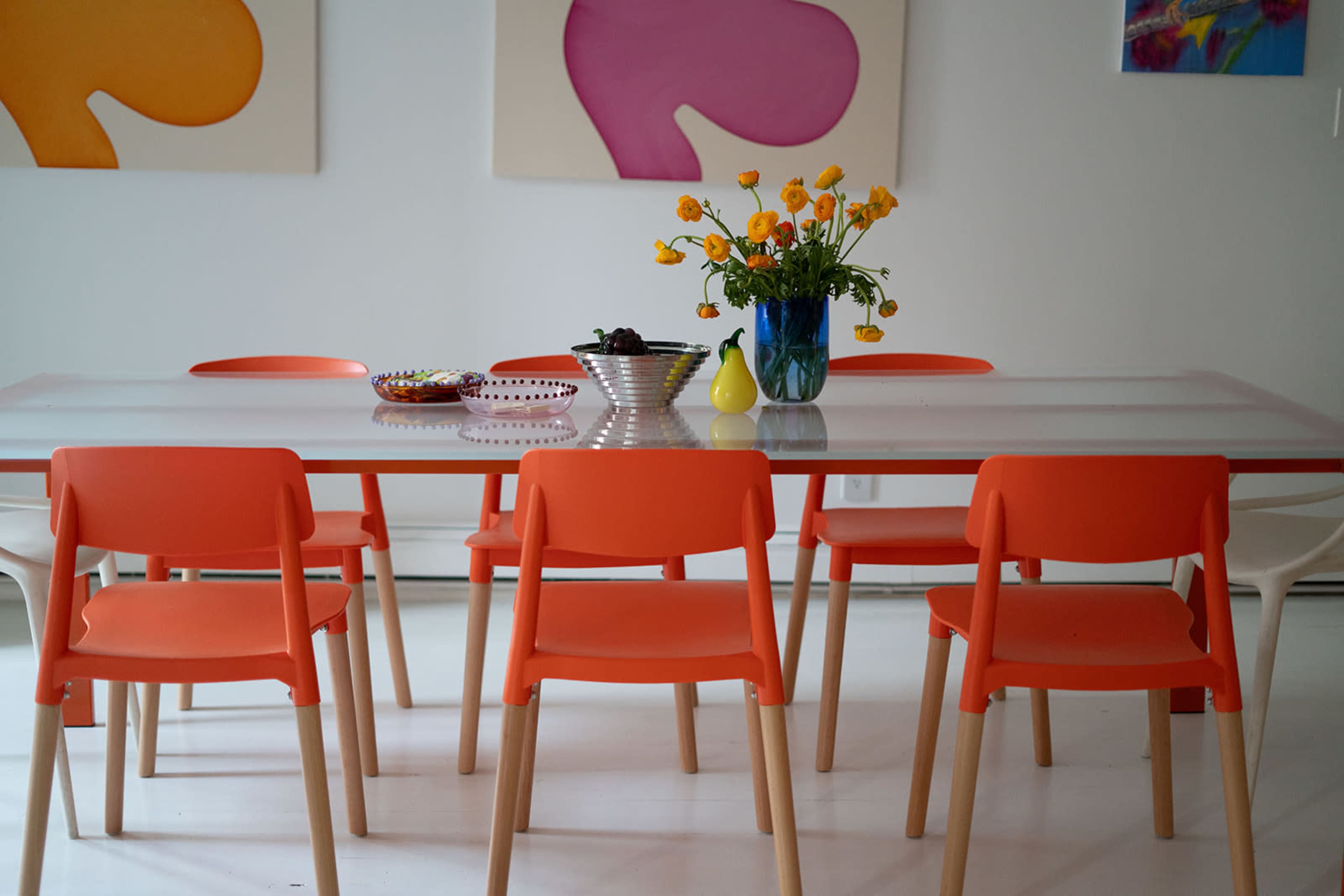 A dining table with a clear top and orange chairs is set with a bowl of fruit, a colorful plate, and a vase of yellow flowers against a backdrop of abstract art.