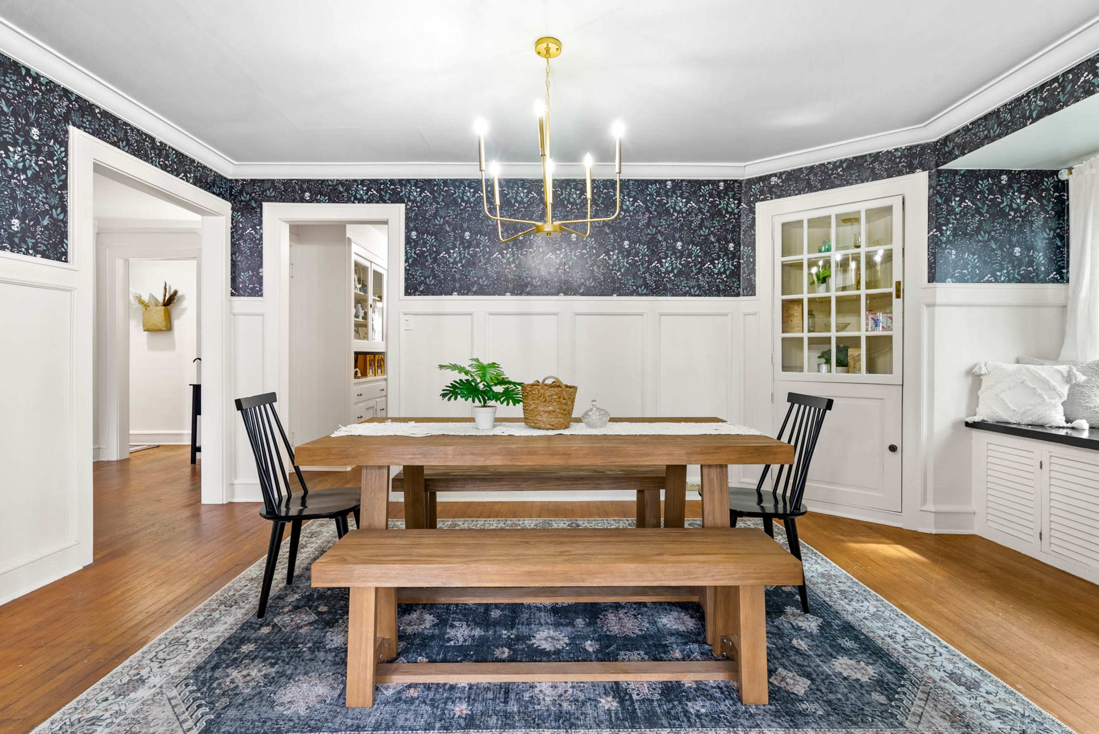 A wooden dining table with a bench is placed in a room featuring floral wallpaper and white wainscoting, illuminated by a modern chandelier.