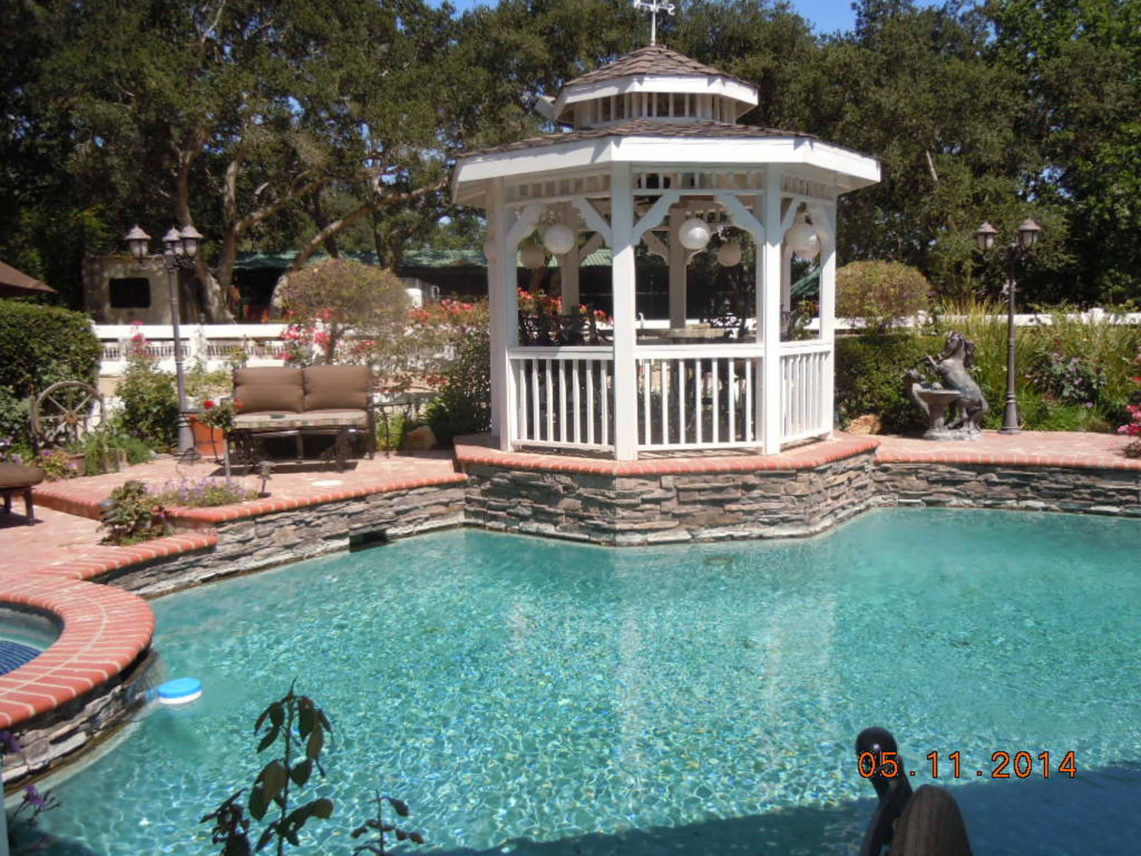 A landscaped pool area featuring a stone gazebo, surrounded by plants and seating.