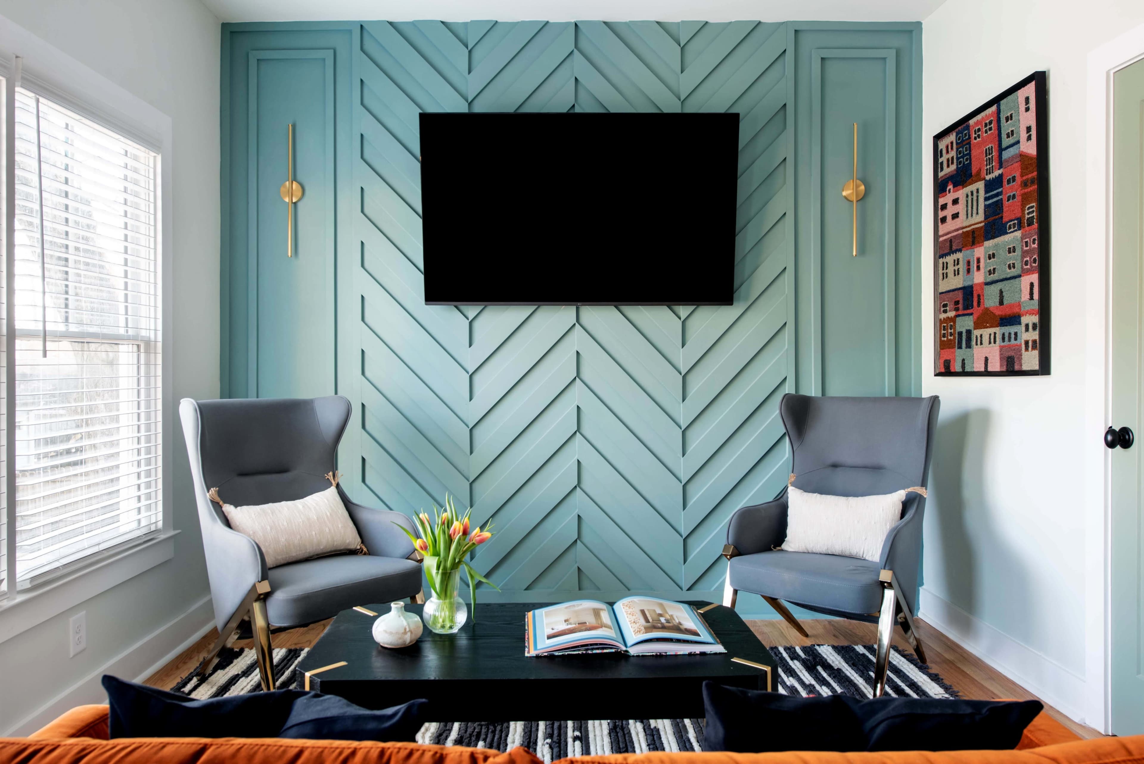 A modern living room with a teal chevron wall, two gray armchairs, a black coffee table, and a flat-screen TV mounted above the table.