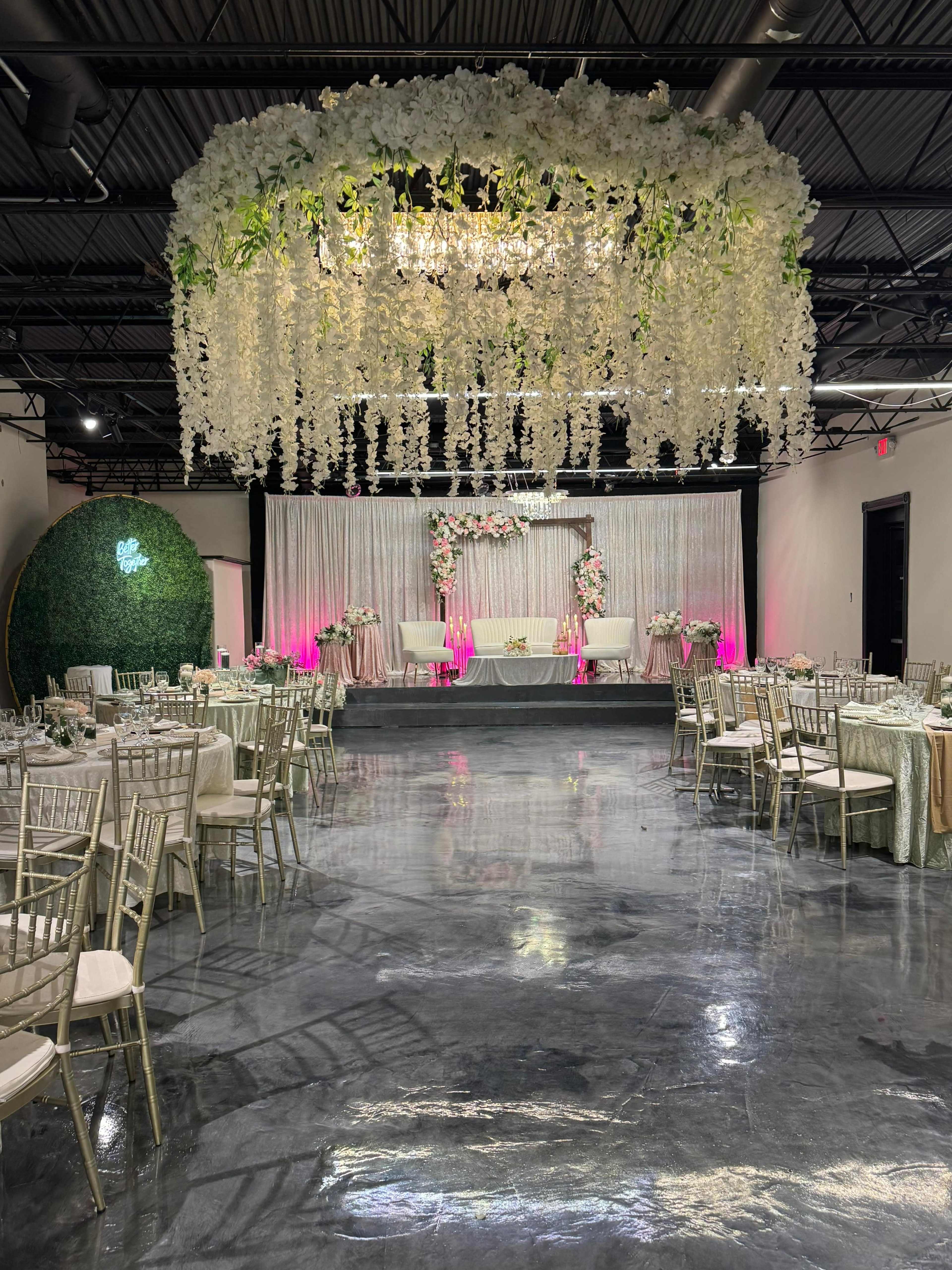 The image shows a decorated event venue with elegantly arranged tables, floral decorations, and a backdrop featuring hanging floral elements and soft lighting.