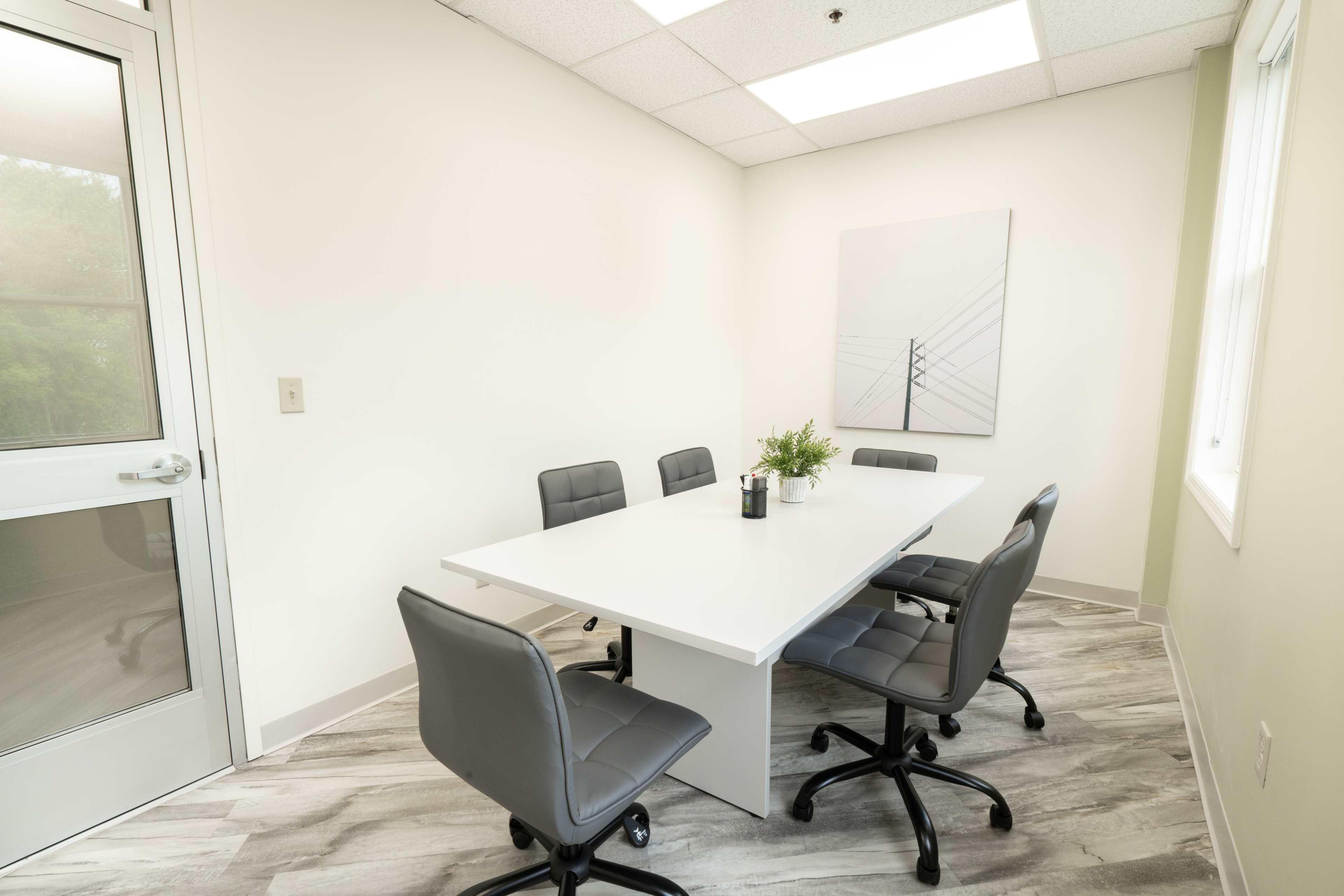 A conference room features a rectangular white table surrounded by five gray office chairs against light-colored walls.