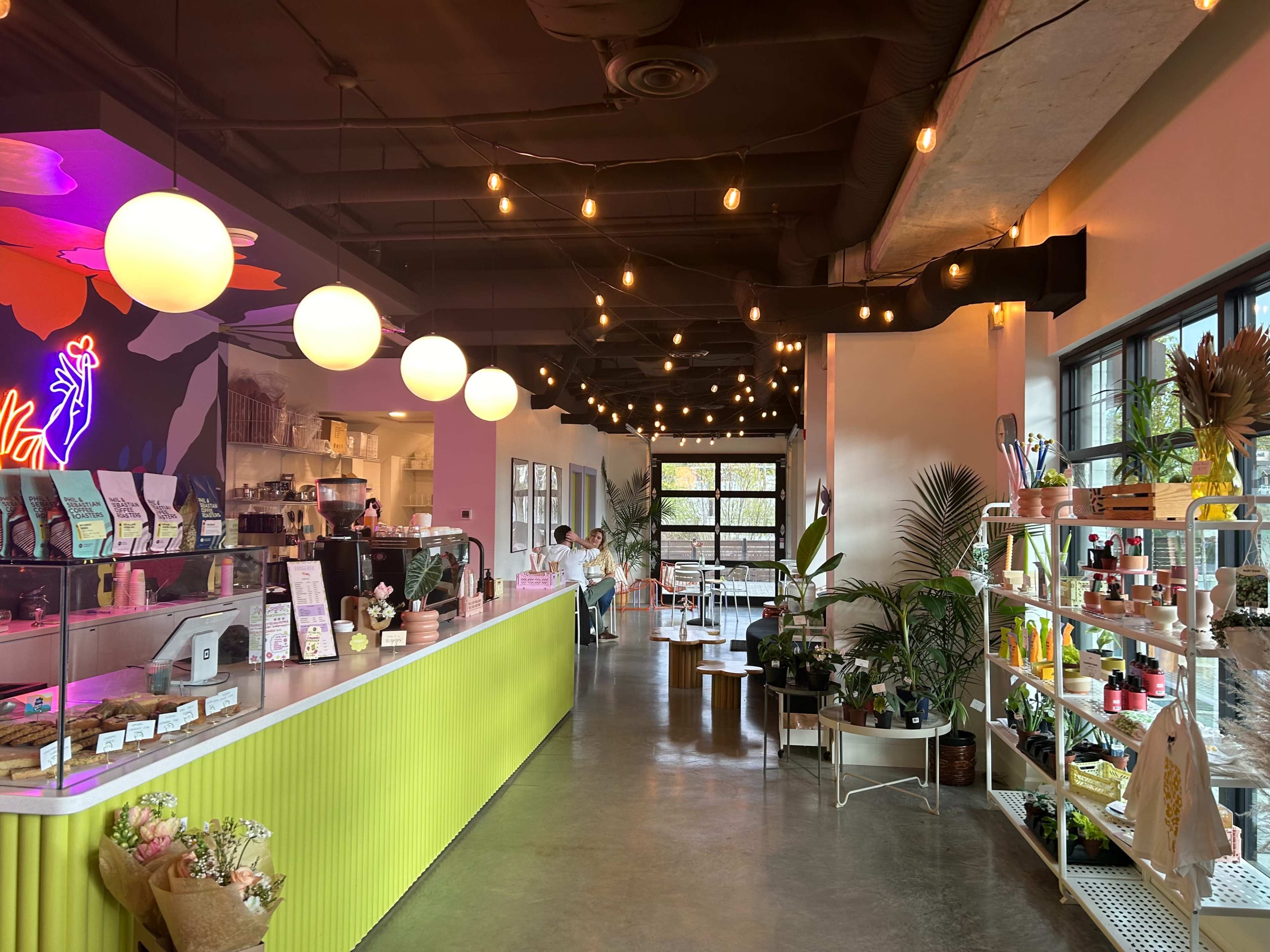 The interior of a modern cafe features a bright green counter, hanging lights, and a display of plants and products for sale, with patrons seated in a cozy setting.