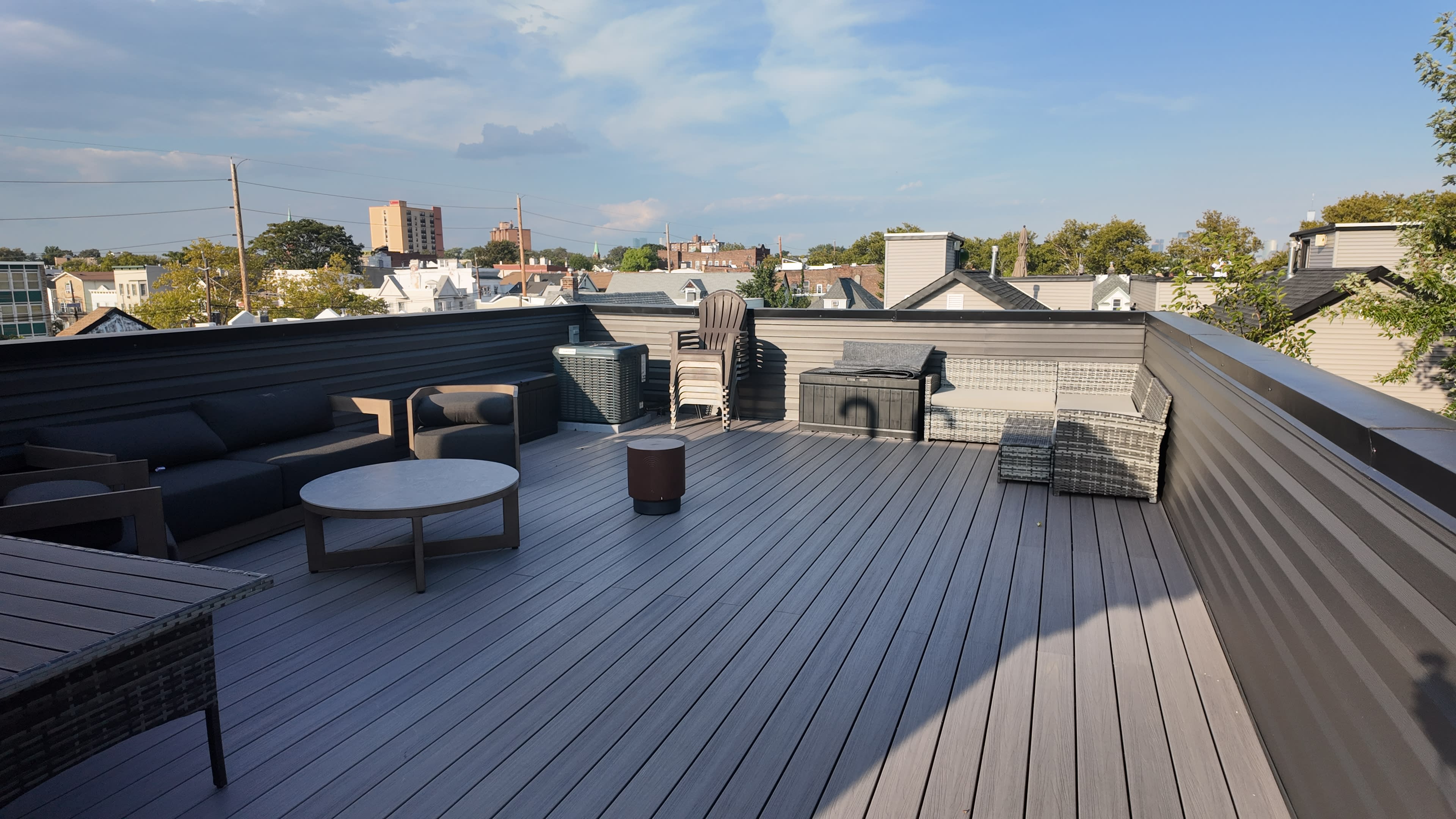 The image shows a flat rooftop deck furnished with outdoor seating, a small table, and a view of city buildings in the background.