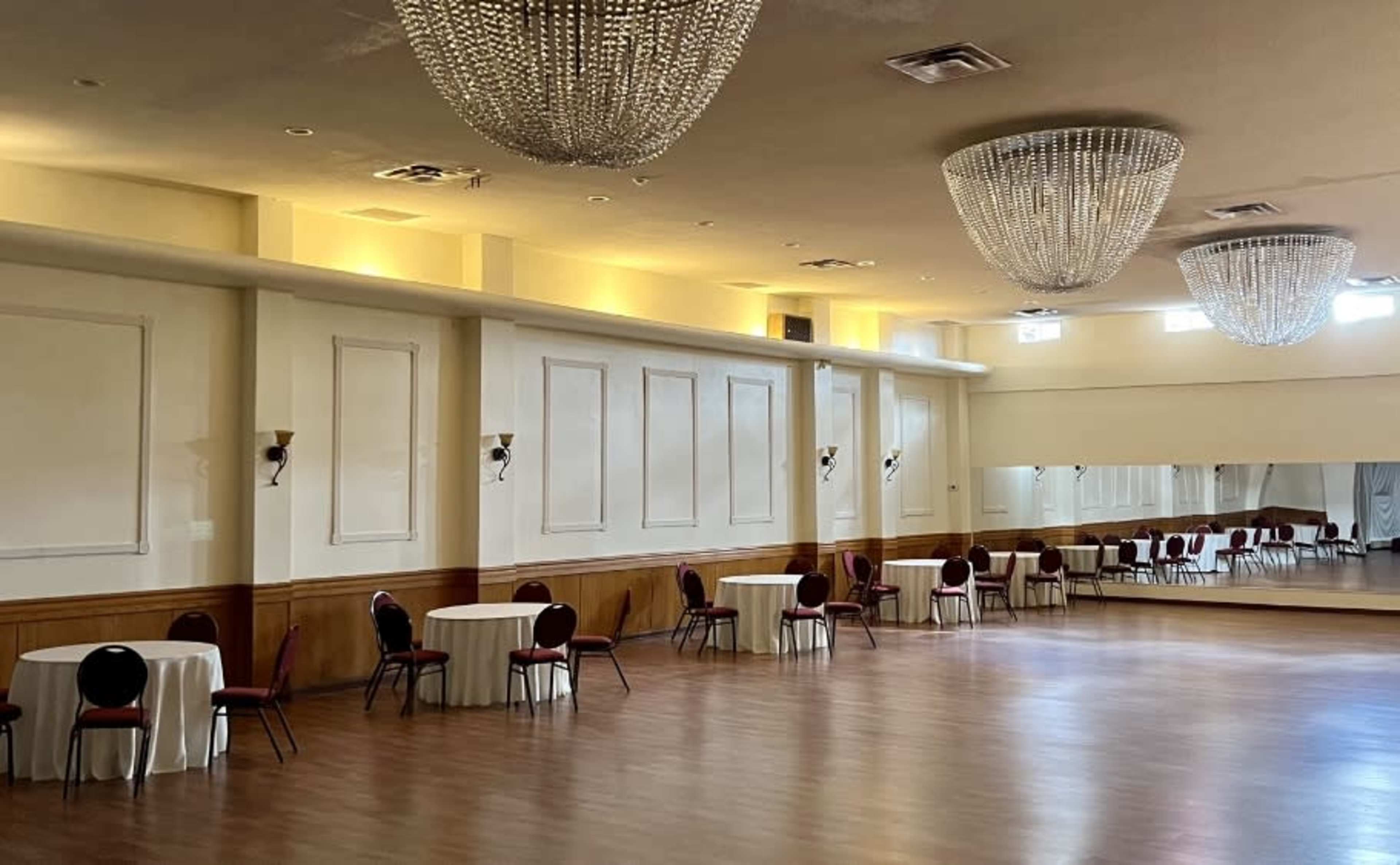 A spacious event hall with round tables and elegant chandeliers, featuring a large mirror on one wall.
