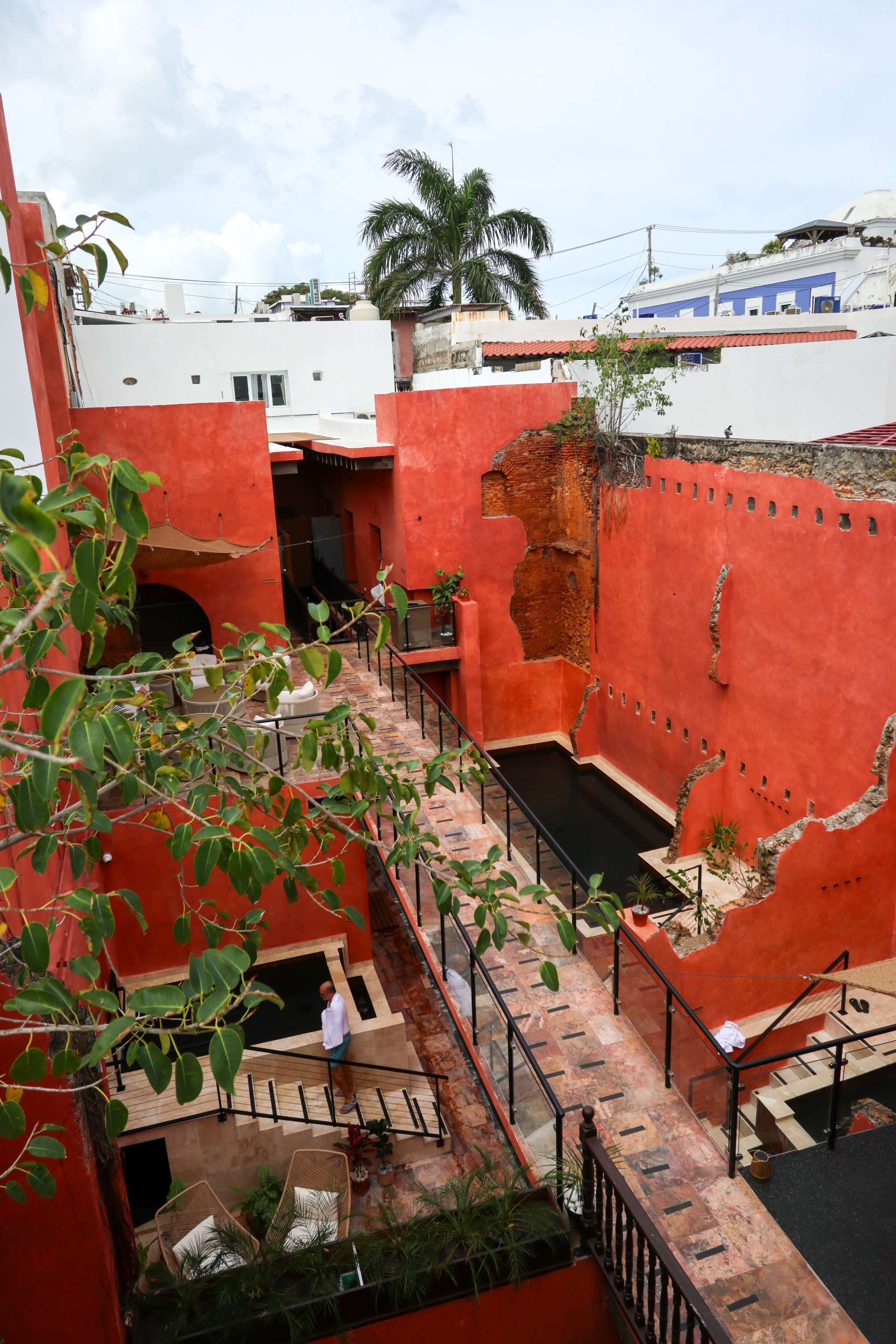 The image shows a courtyard with red walls, featuring a pathway flanked by plants and water features, with several people walking around.