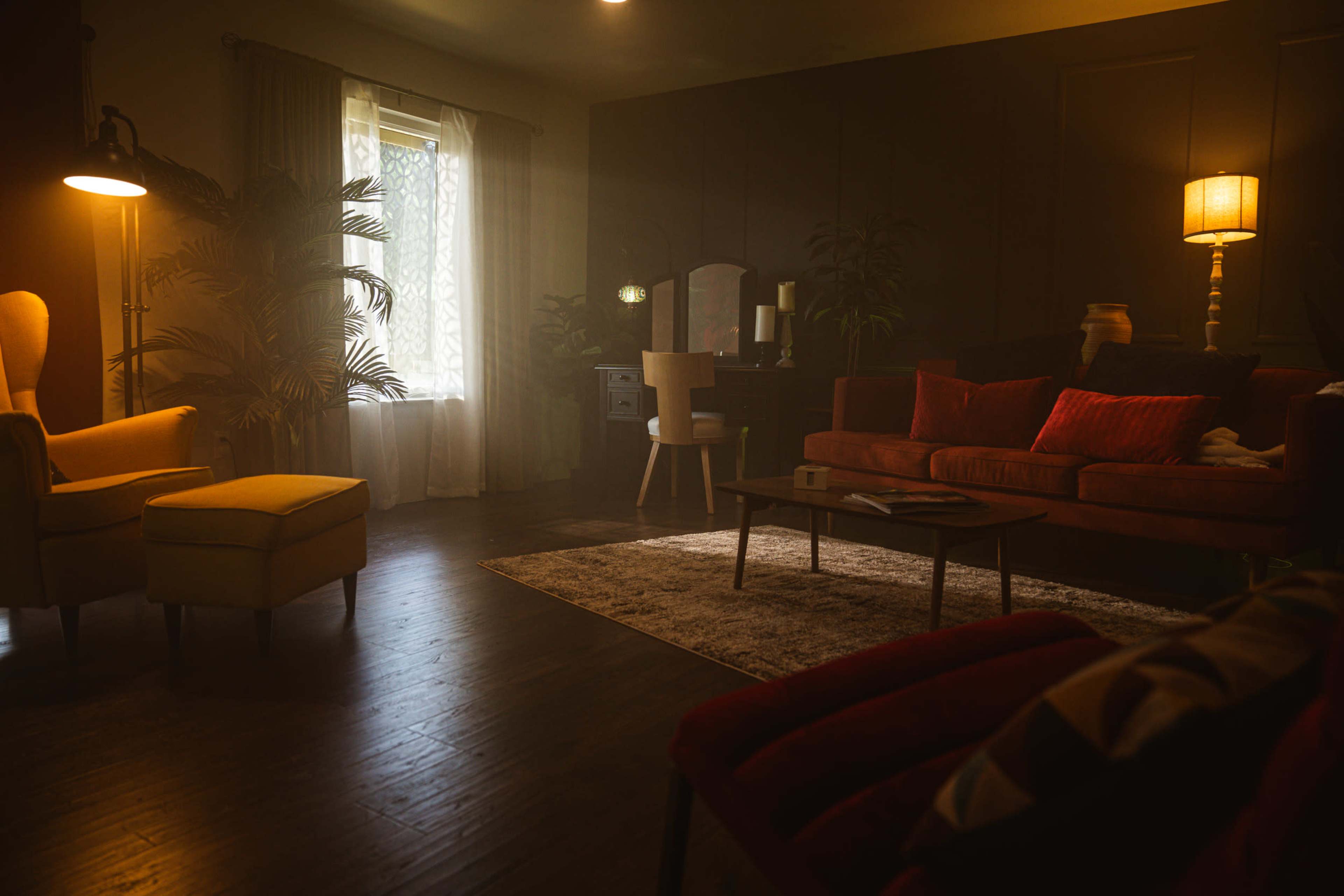 A dimly lit living room features a sofa, armchairs, a coffee table, and a window with sheer curtains.