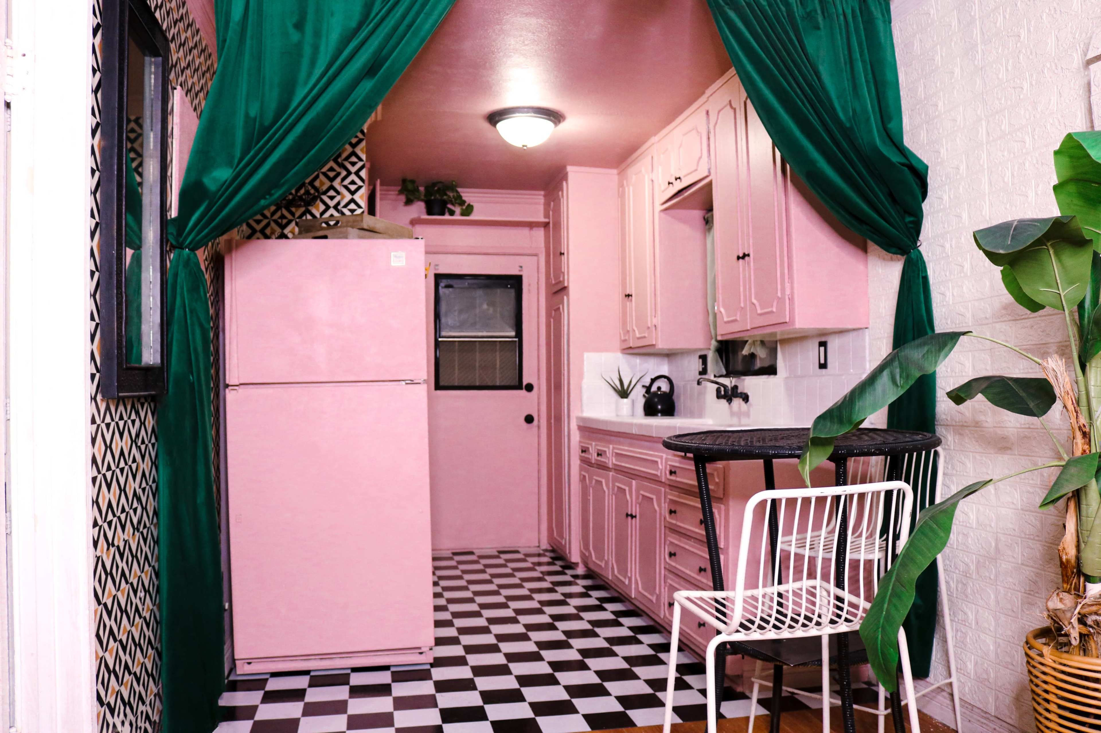 The image shows a narrow kitchen featuring pink cabinets and a refrigerator, black and white checkered flooring, and curtains draping from the ceiling.