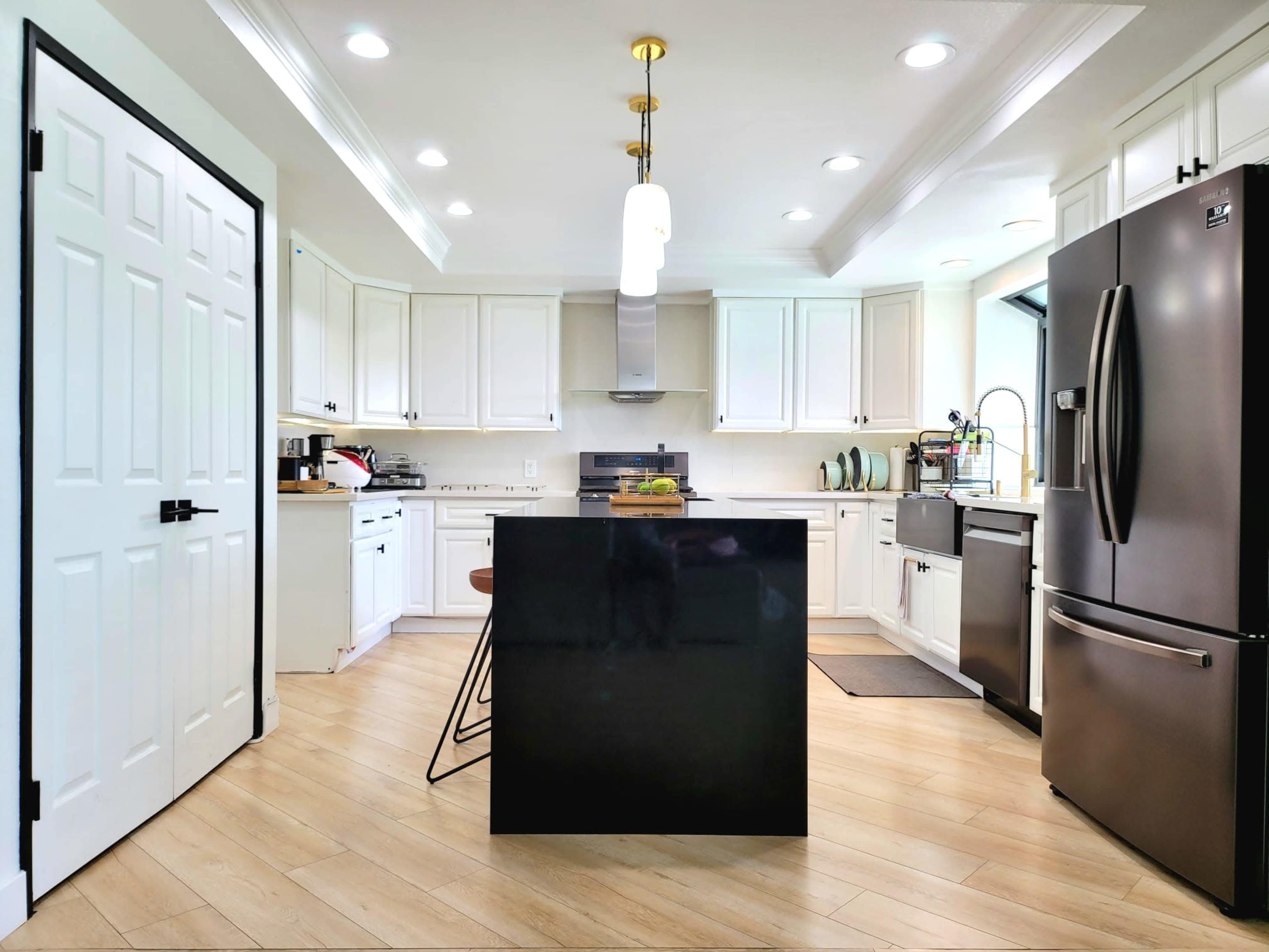 A modern kitchen with white cabinets, a black island, stainless steel appliances, and wooden flooring.