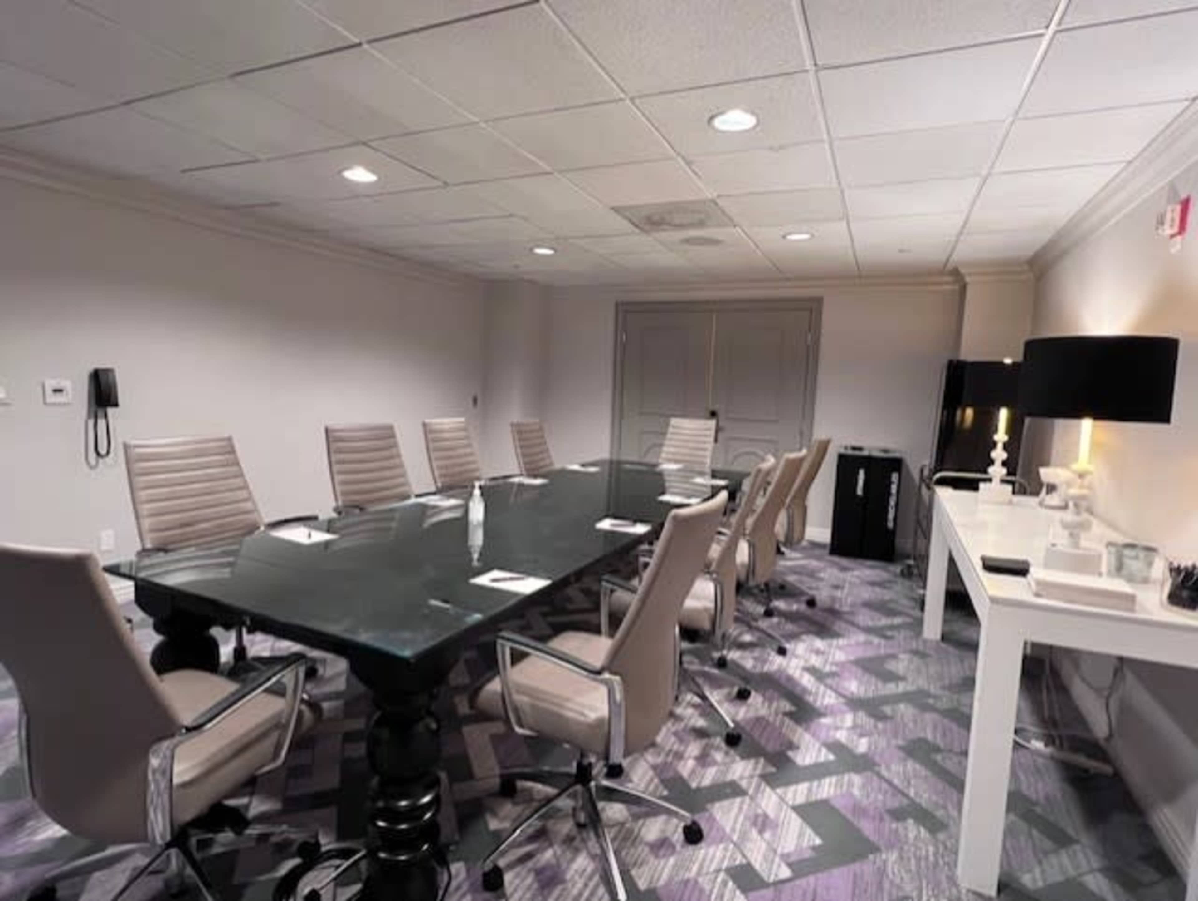The image shows a modern conference room with a large glass table, eight ergonomic chairs, and additional furnishings including a side table and a lamp.