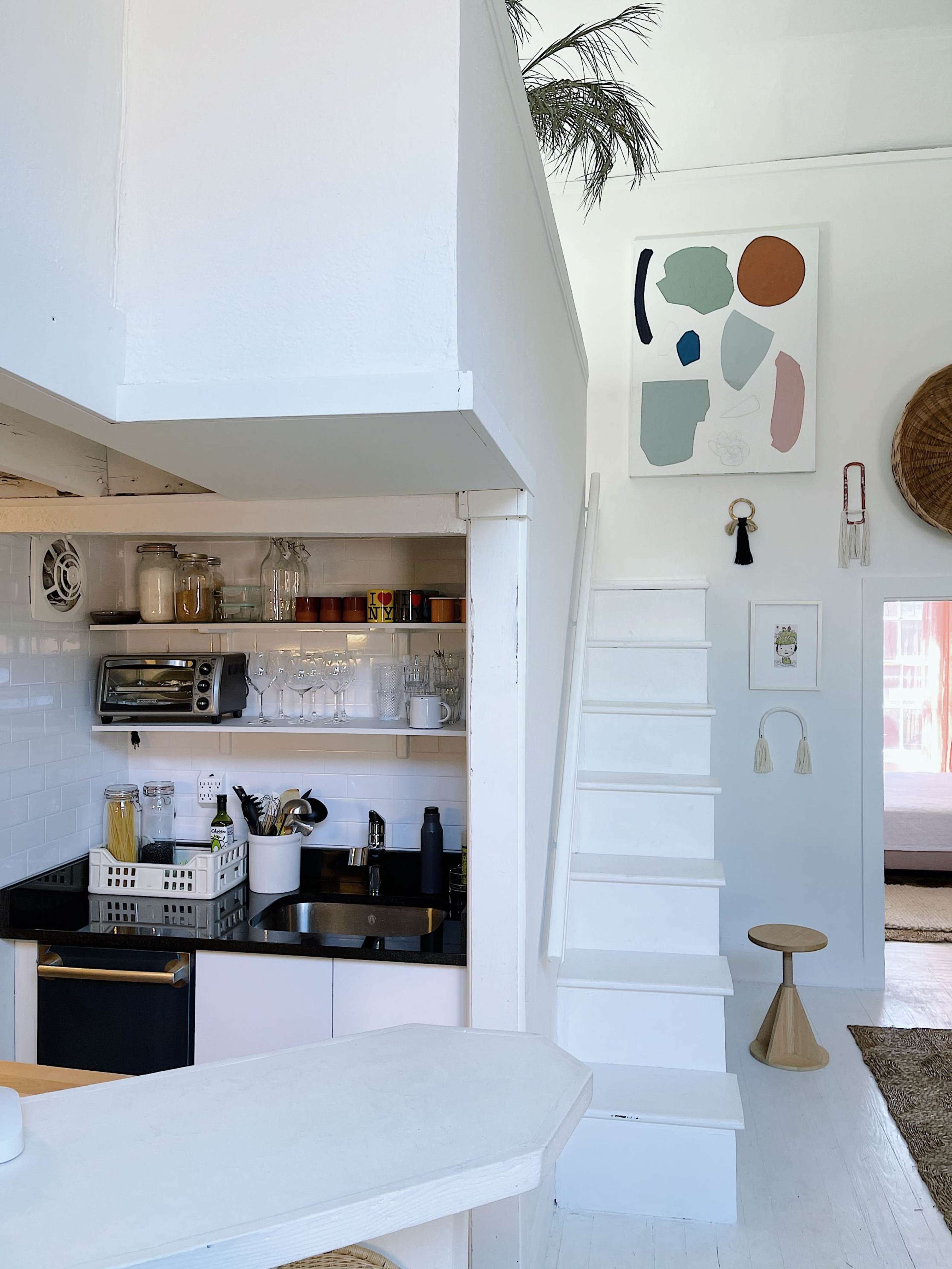 A modern kitchen with open shelving, featuring a black countertop and a staircase leading to an upper level with artwork on the wall.