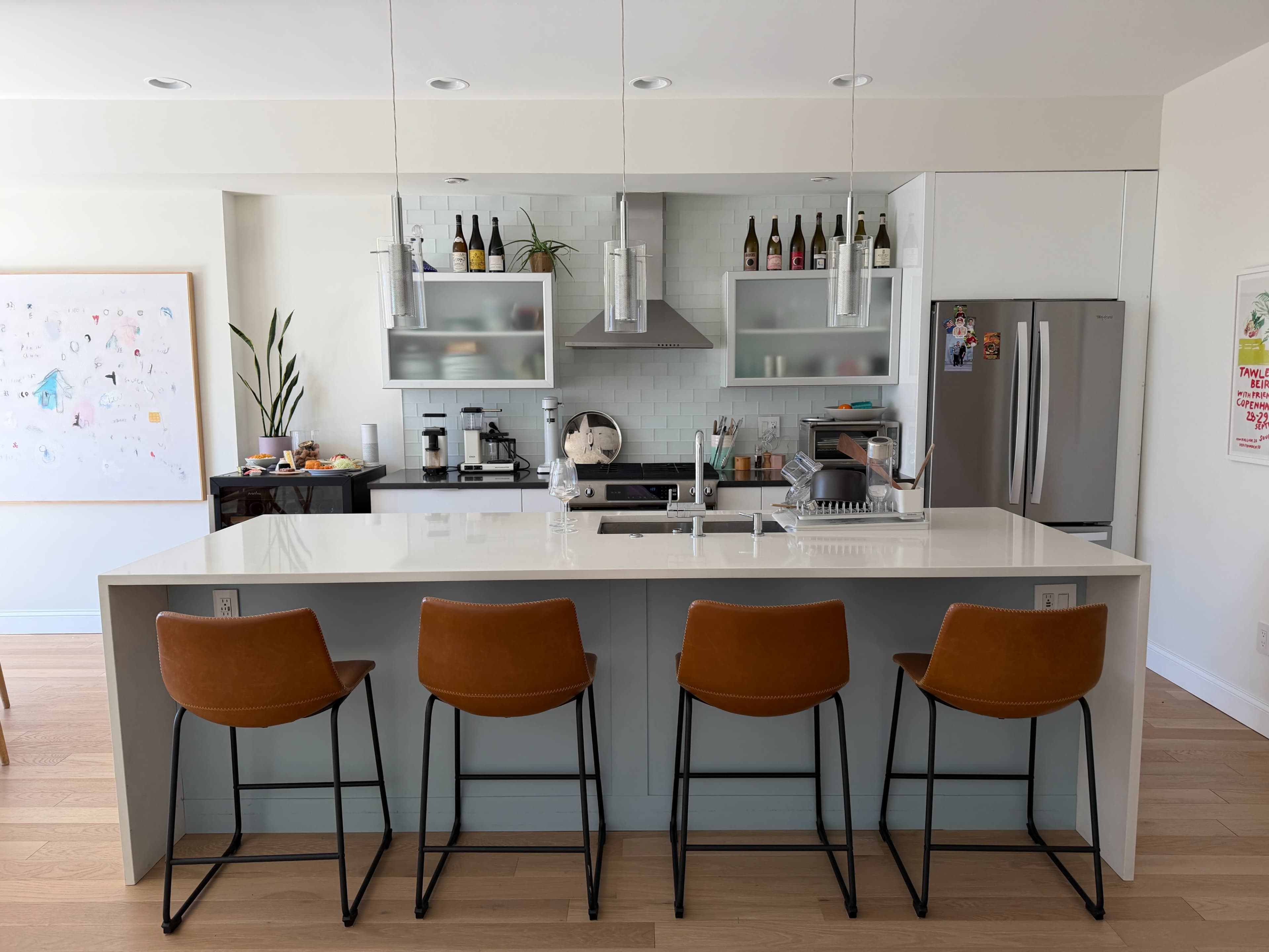 A modern kitchen features a central island with four brown bar stools, sleek cabinetry, and a variety of kitchen appliances and decorative items in the background.