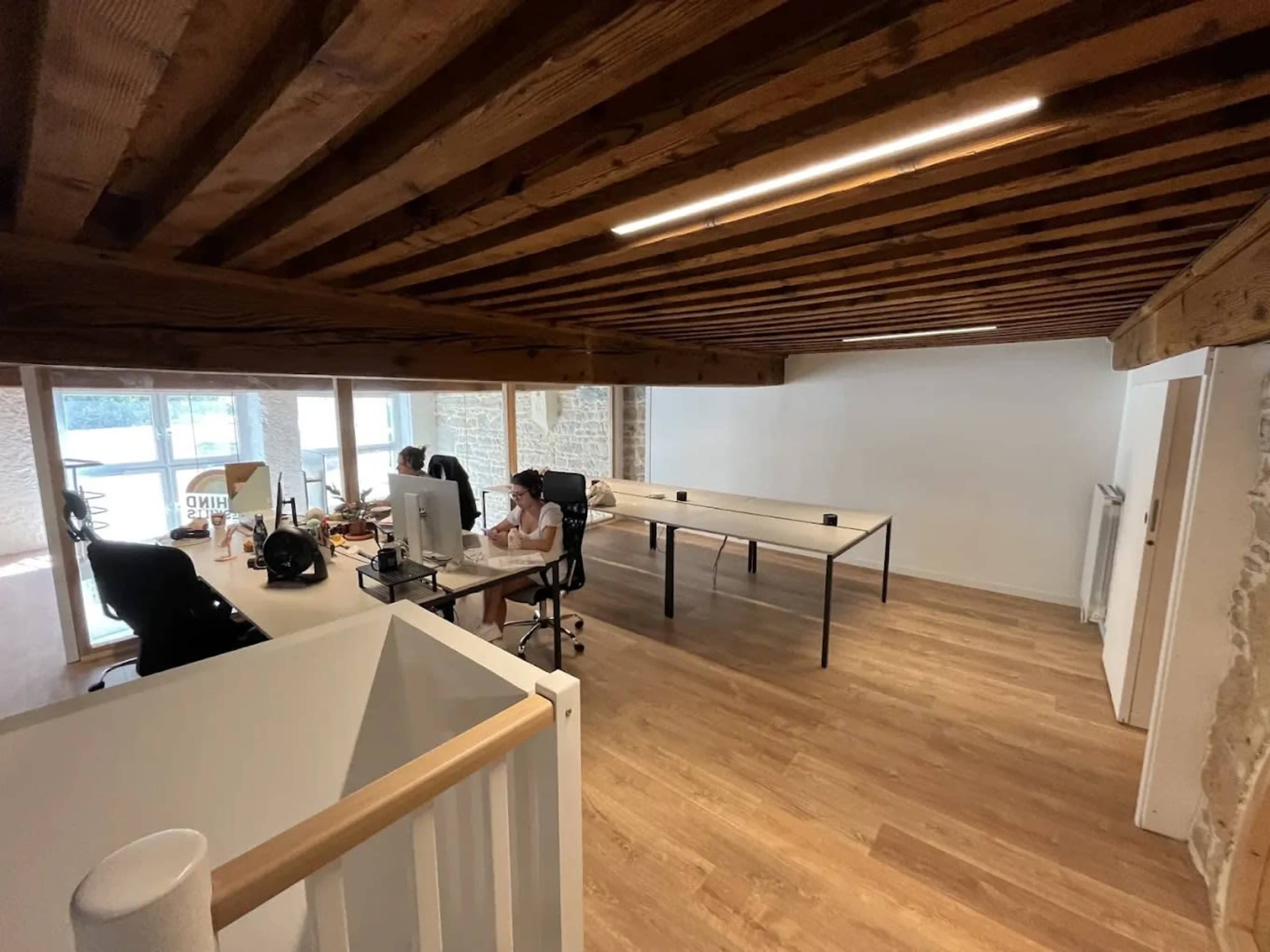 The image shows an office space with wooden beams, a large desk, two workstations, and ample natural light from large windows.
