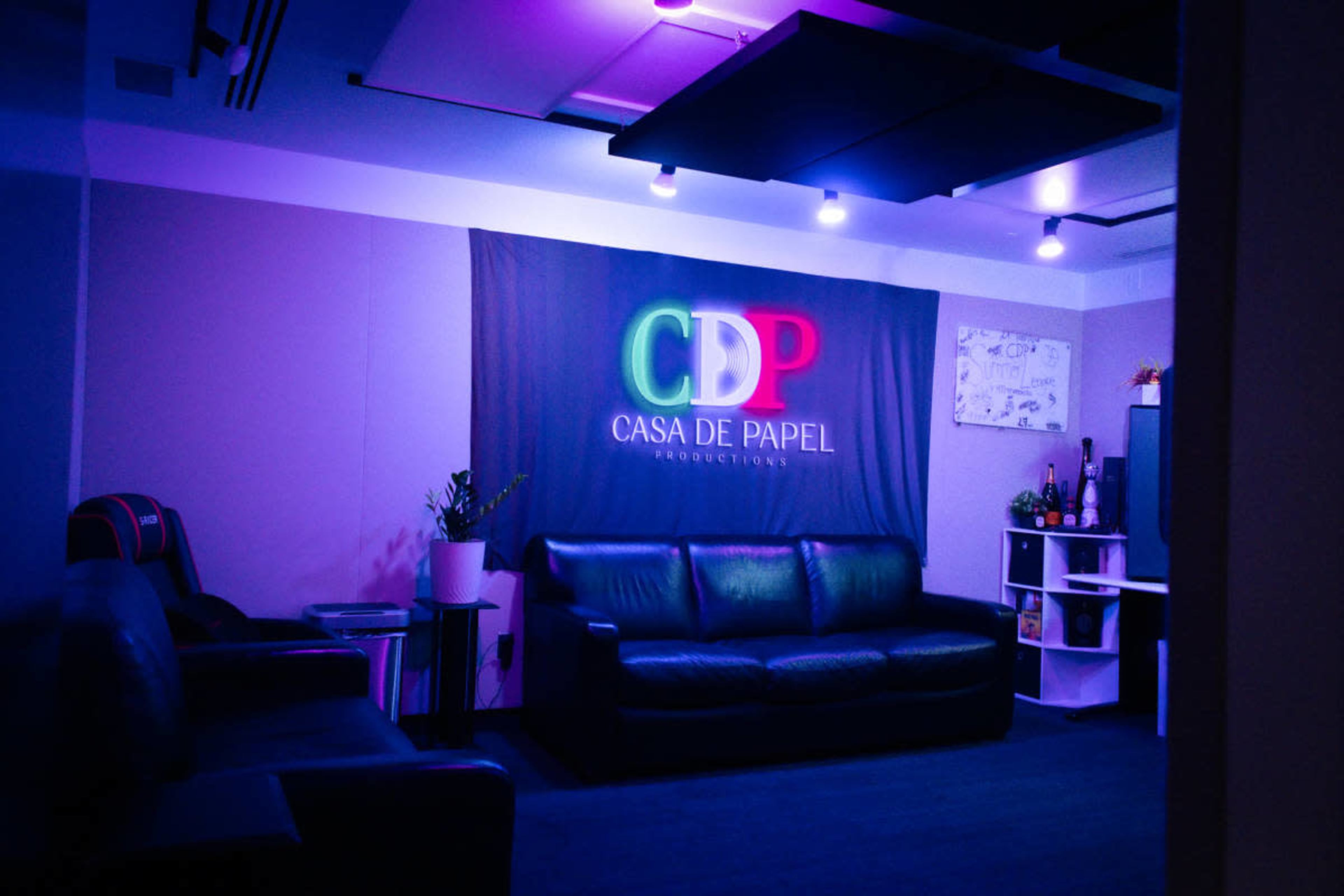 The image shows a dimly lit room featuring a black leather sofa, a decorative wall with colorful lighting displaying "CDP CASA DE PAPEL PRODUCTIONS," and various furniture items.