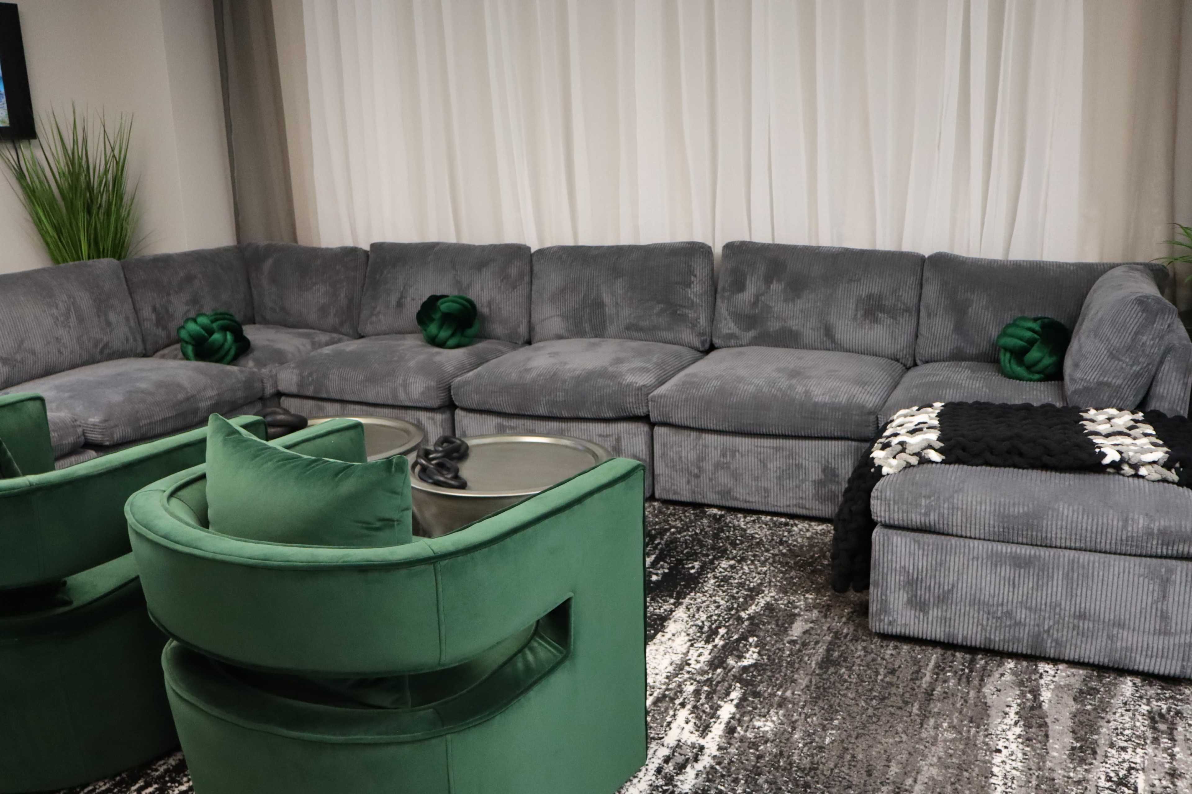 A spacious living room featuring a grey sectional sofa, two green accent chairs, and a dark patterned rug.