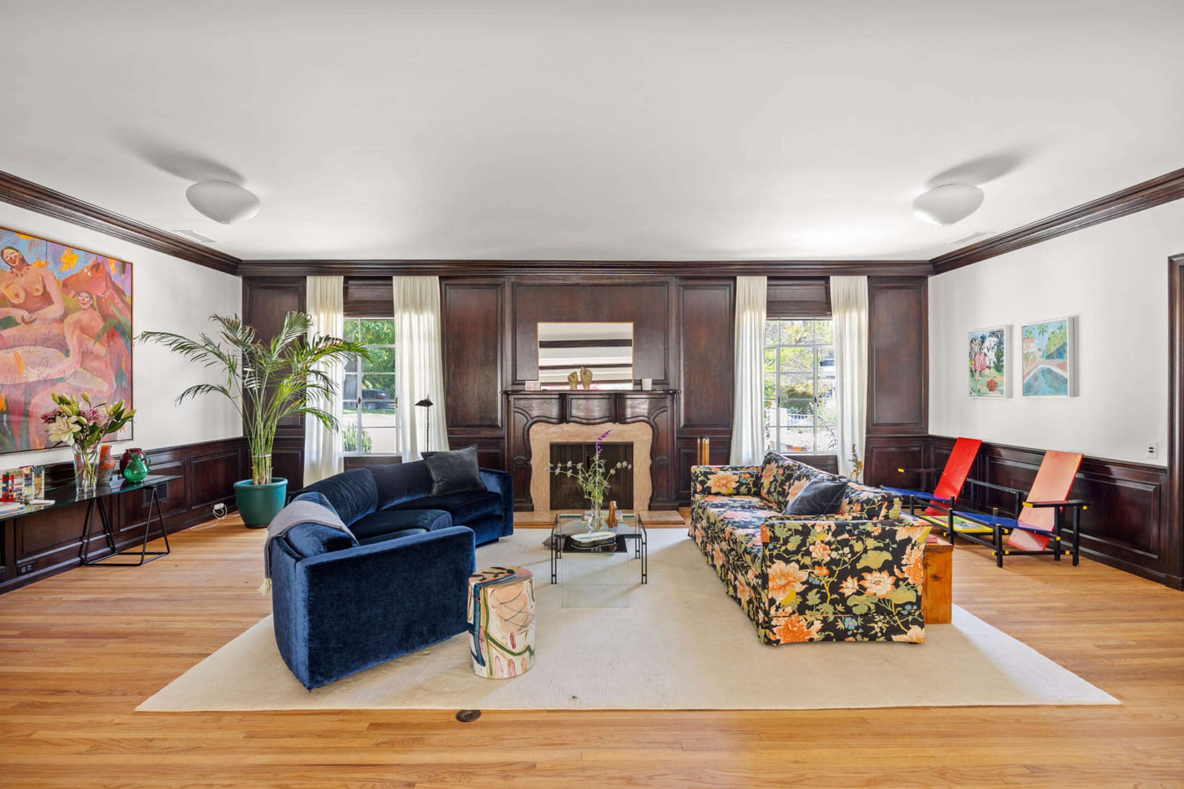The living room features a mix of dark wood paneling, two distinct sofas—one blue velvet and one floral, a coffee table, and large windows letting in natural light.
