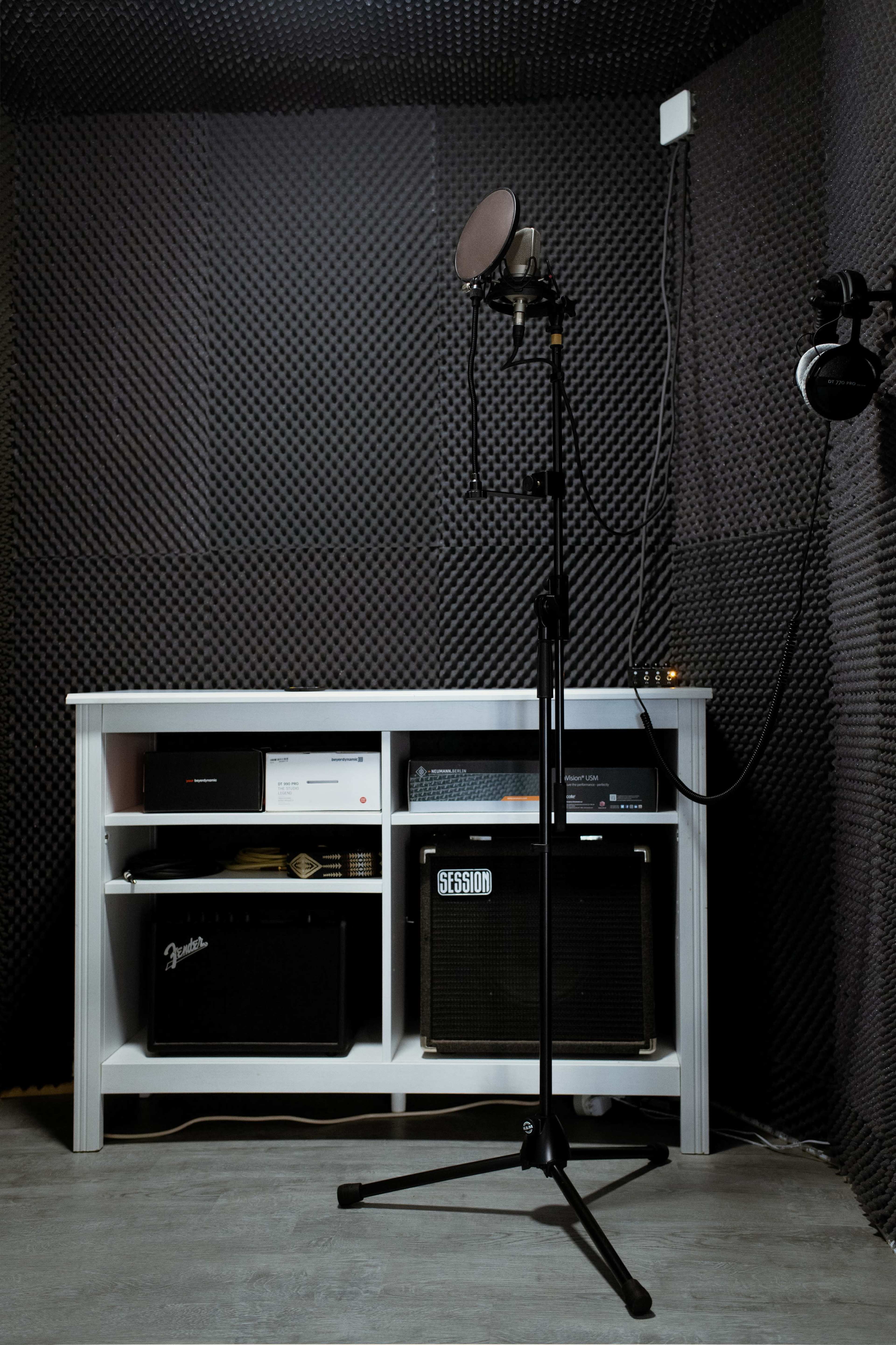 A microphone stand is positioned in front of a white shelf filled with audio equipment, all set within a soundproof room lined with black acoustic panels.