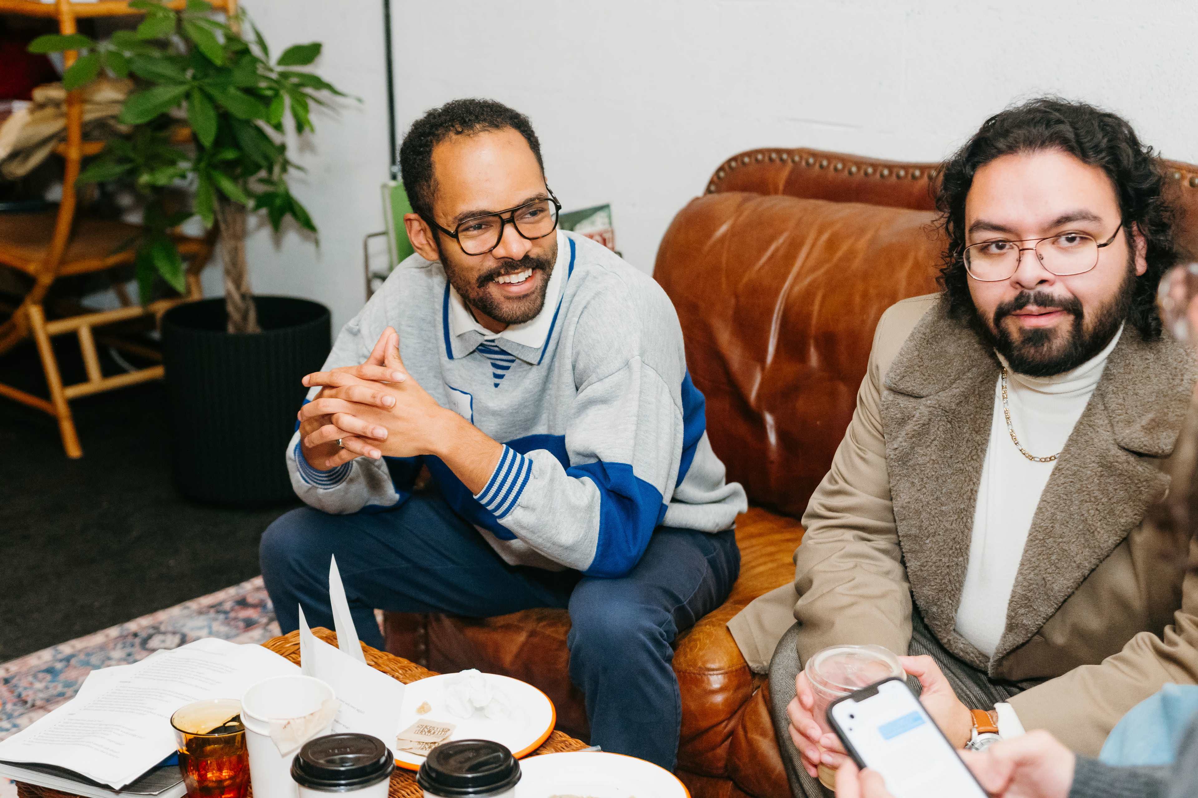 Two men are seated on a brown leather couch in a casual setting, engaged in conversation, with plates and beverages on a coffee table in front of them.
