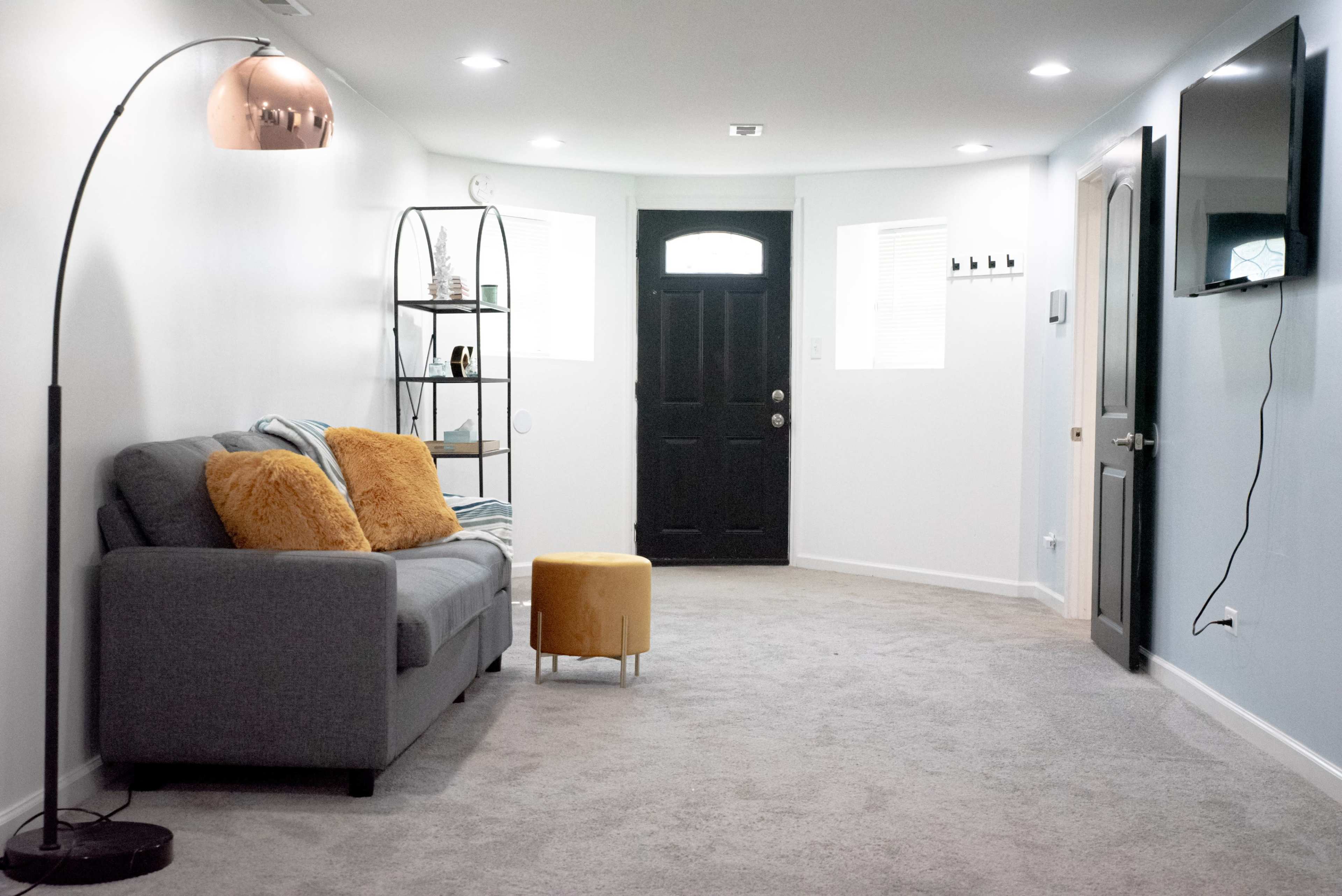 A modern living space with a gray sofa, a yellow ottoman, a floor lamp, and a black door, all set against light gray walls and carpet.