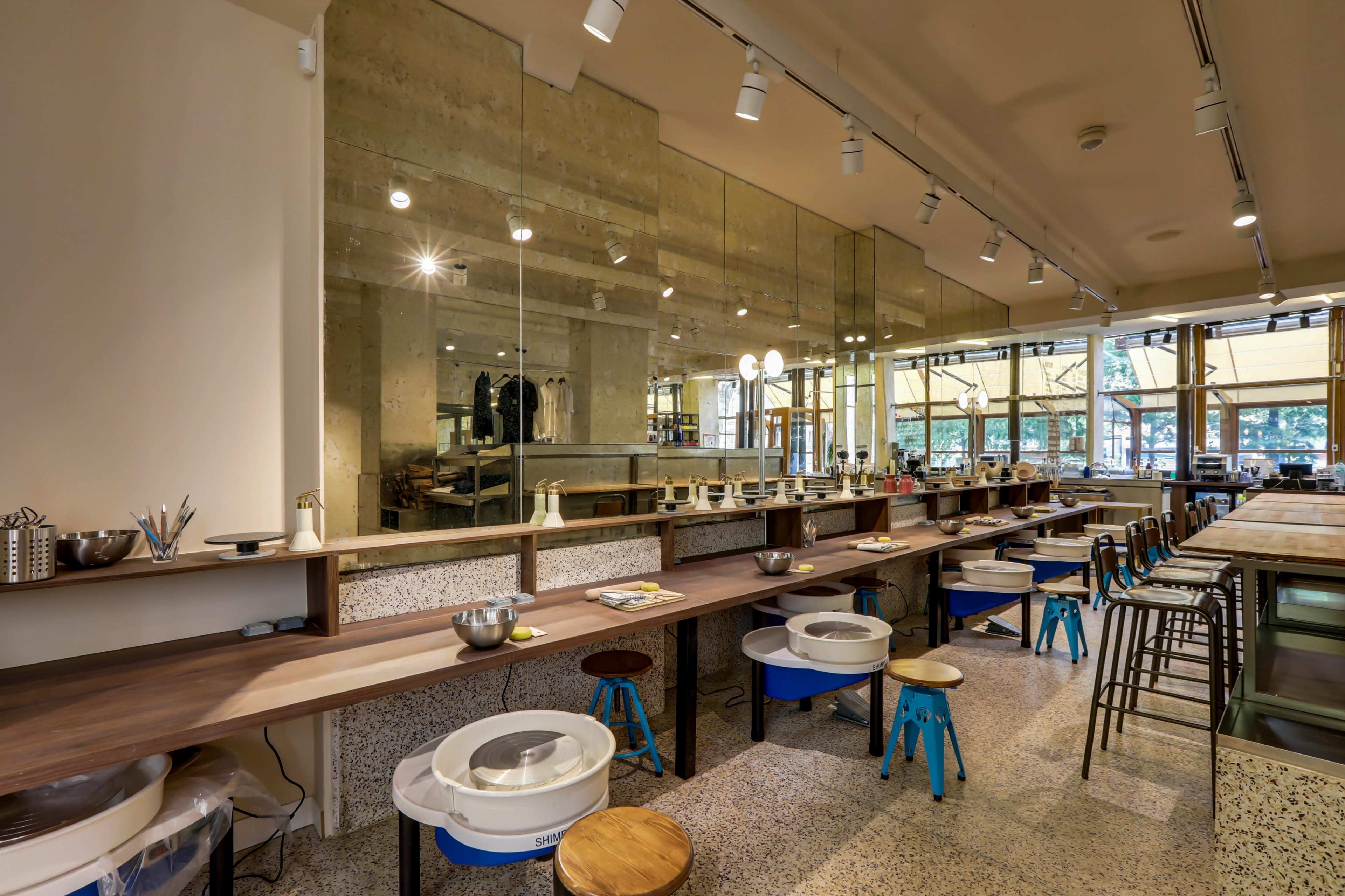 The image shows a modern pottery studio with workstations featuring pottery wheels, tools, and a large mirror reflecting the interior space.