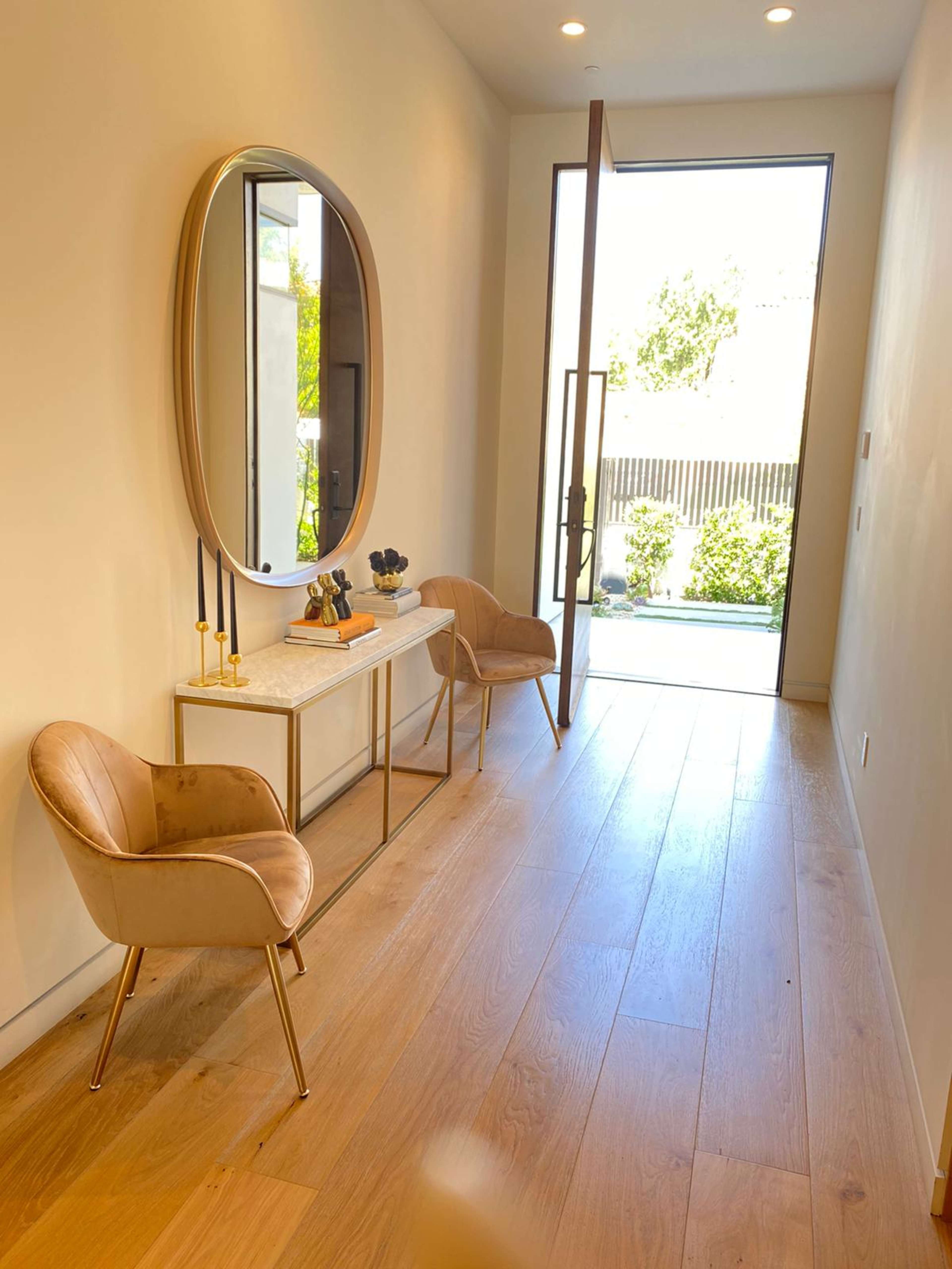 A modern entryway with two stylish chairs, a console table, and a large round mirror, leading to a glass door that opens to a garden.