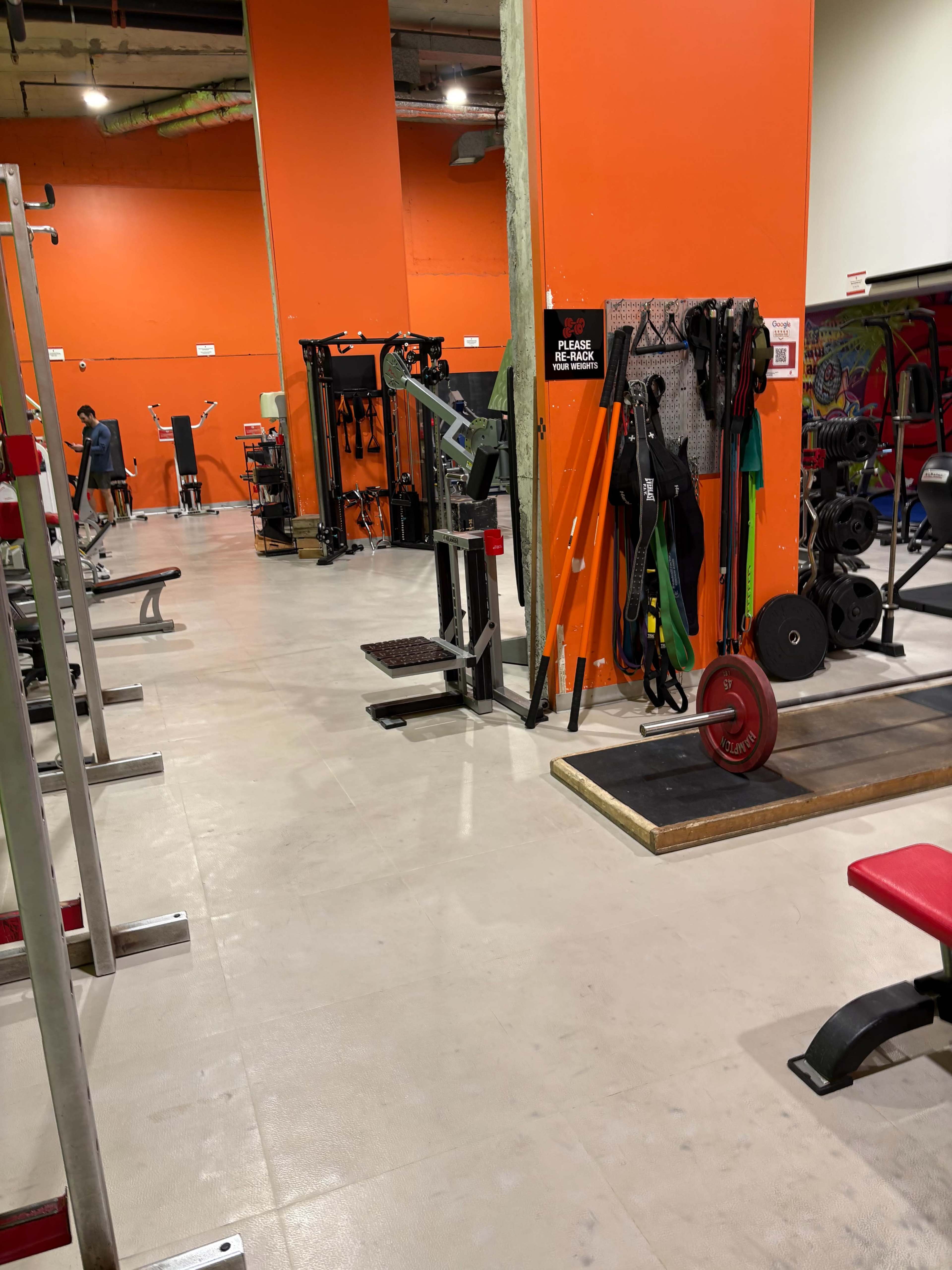 The image shows a gym interior with orange walls, featuring various weightlifting and exercise equipment arranged neatly along the floor.
