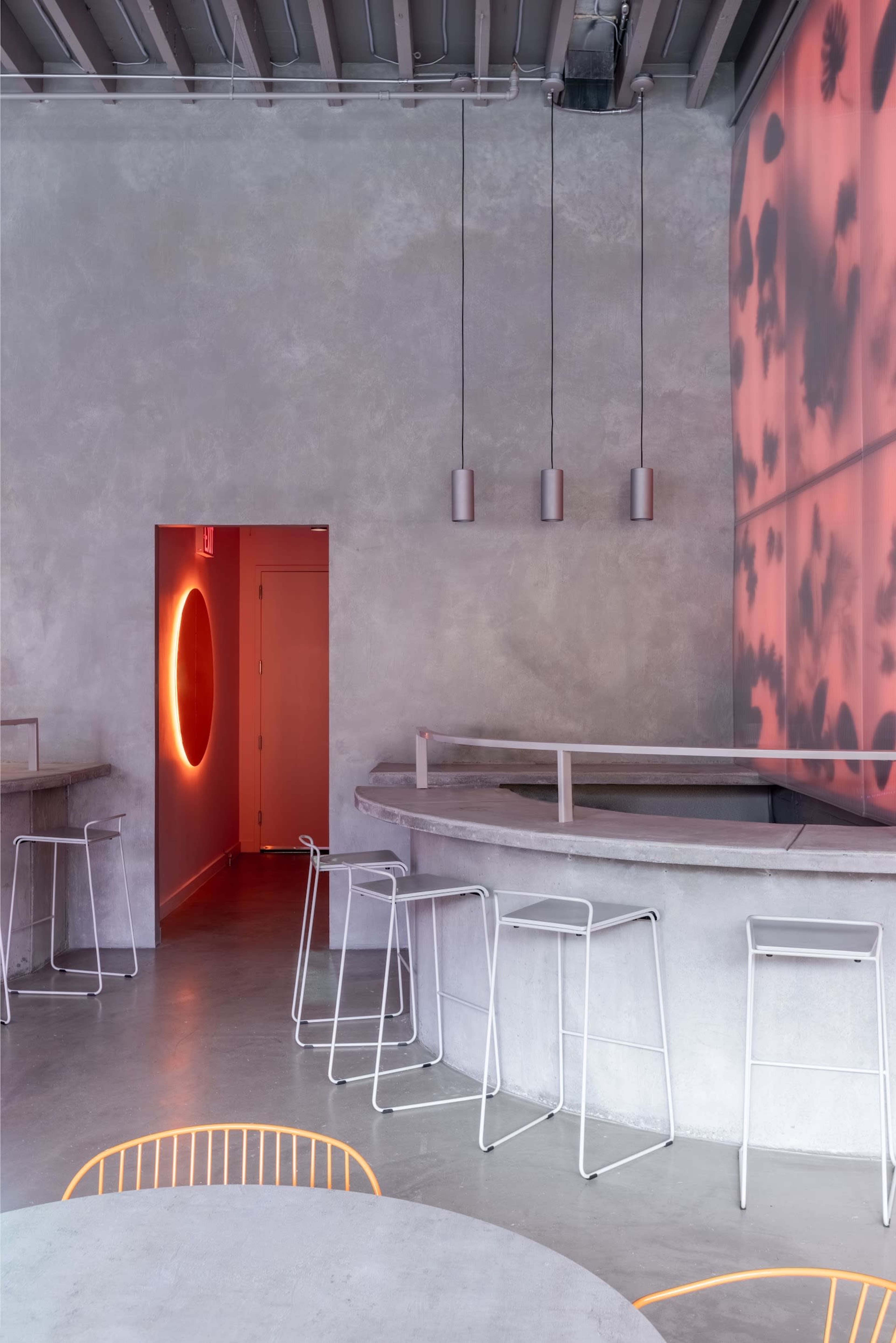 A modern, minimalist café interior featuring a curved counter, several high stools, and a doorway illuminated with a circular light.