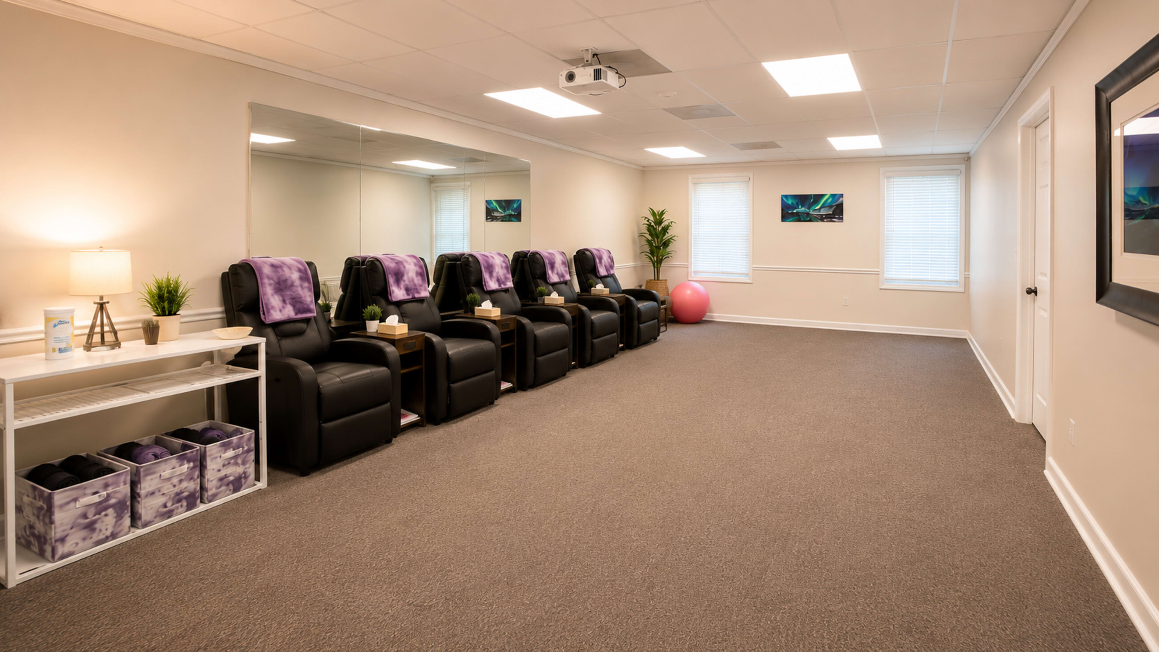 The image shows a spacious room with six black reclining chairs, a large mirror, and a soft pink exercise ball, illuminated by natural light from windows.