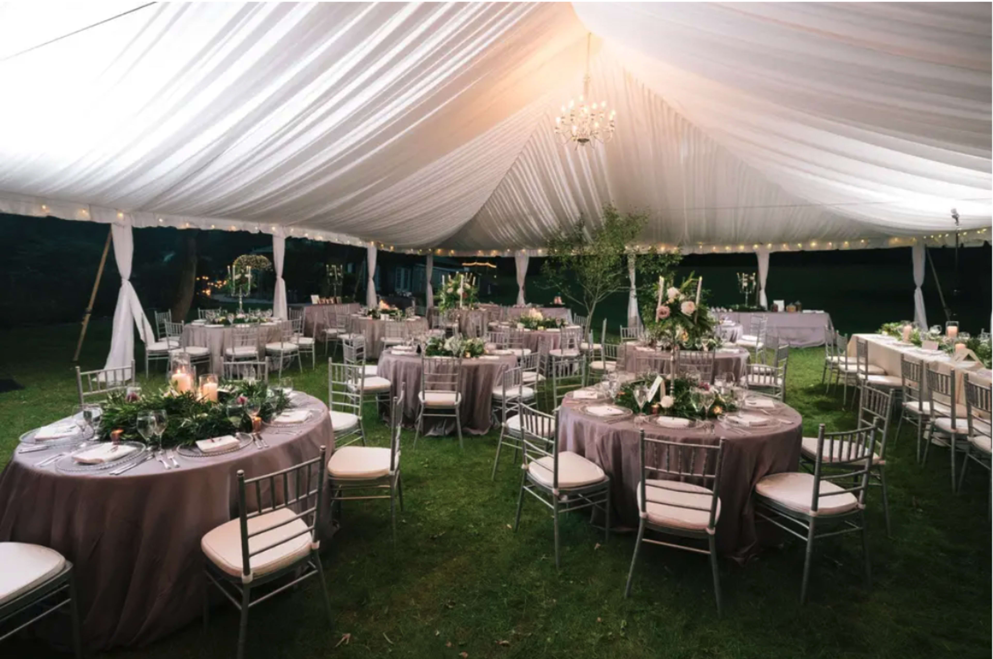 A large outdoor tent is set up for an event, featuring round tables with gray tablecloths and silver chairs, all decorated with floral centerpieces and candles.